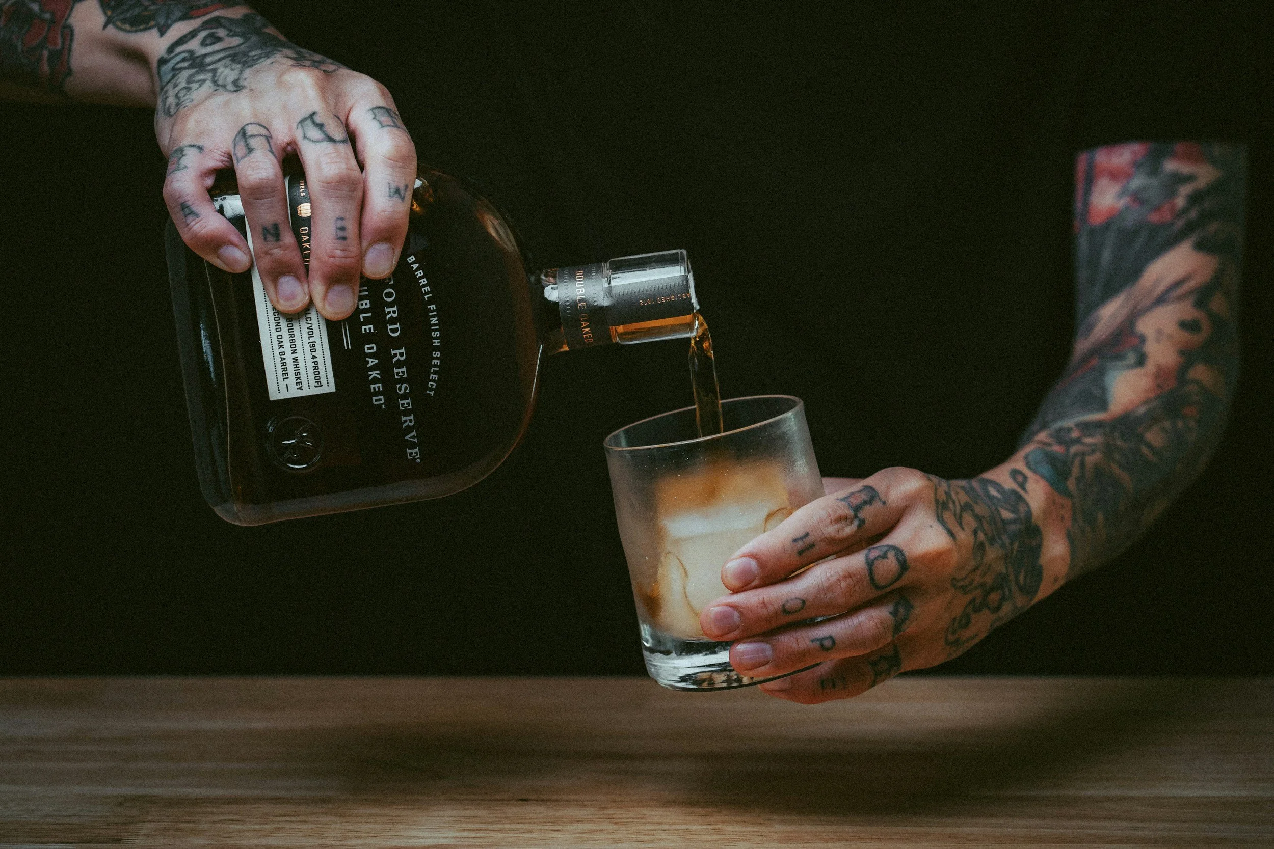 A person with tattooed hands pouring liquor into a glass of ice on a dark background.