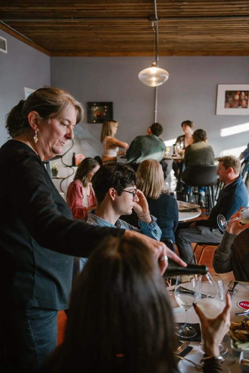A group of people dining and socializing in a restaurant during evening, with some drinking and others engaged in conversation.
