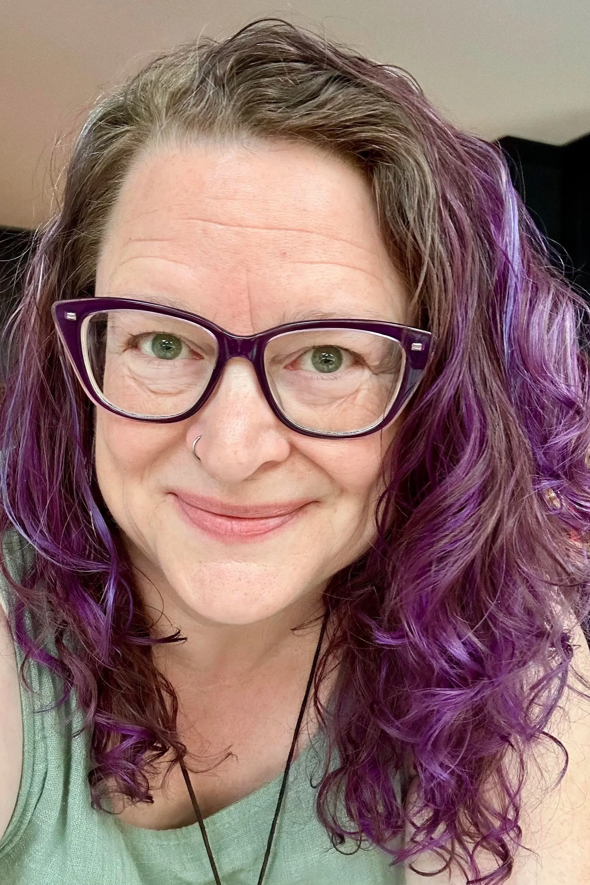 A woman with curly hair that is brown at the roots and purple towards the ends, wearing purple glasses and a light green top, smiling at the camera.