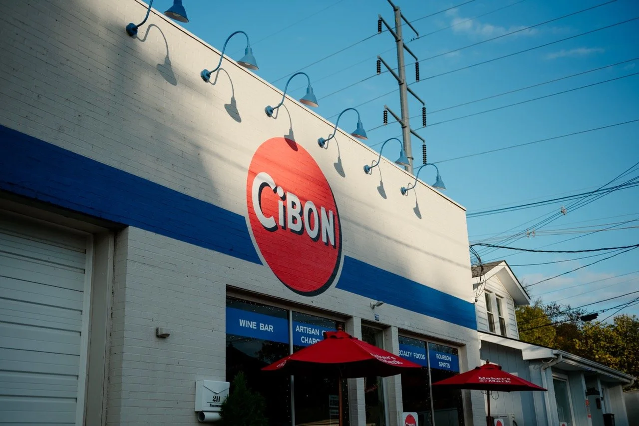 Exterior of a white brick building with a large red circle logo displaying "CIBON" in the center. There are blue awning lights above and two red umbrellas outside, indicating an outdoor seating area. The building has large windows with signage advertising wine bar, artisan cheese, and spirits. Power lines and a small white house are visible in the background under a clear blue sky.