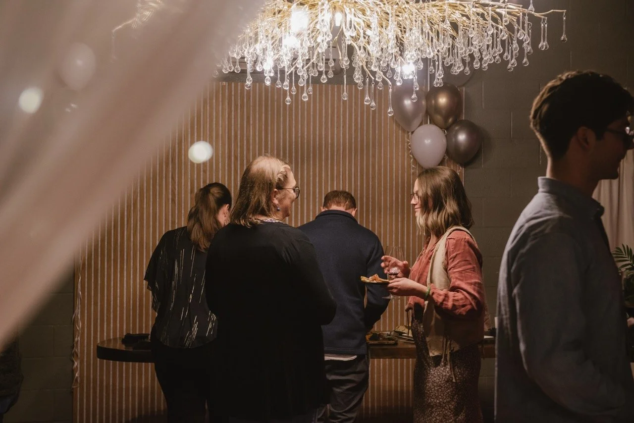 People socializing at a party or gathering, with a chandelier overhead, balloons in the background, and a wooden wall.