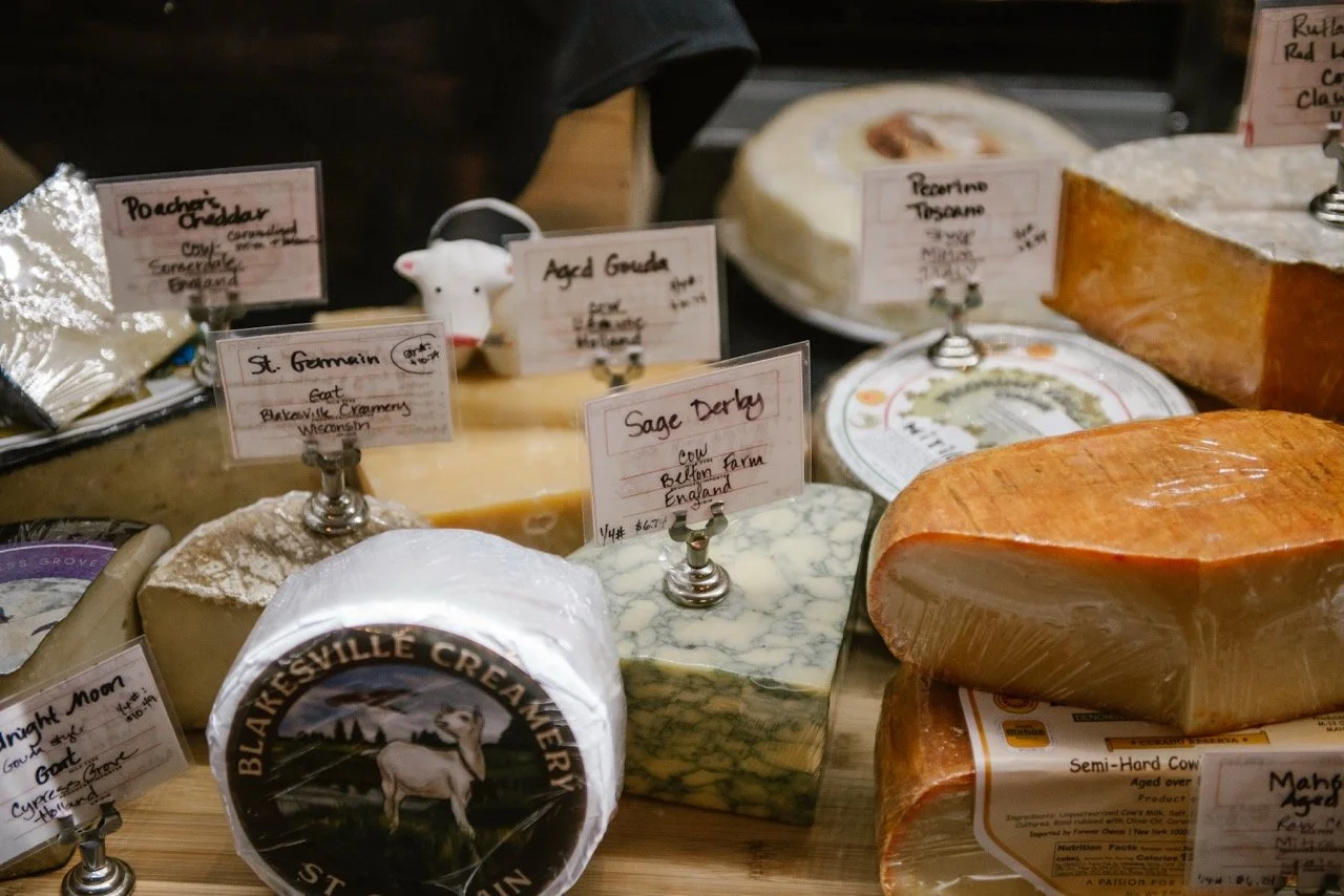 Assorted cheeses displayed at a cheese shop with labels including Blaesville Creamery, St. Germain, Sage Derby, Rocchette Toscano, and semi-hard cow's milk cheese.