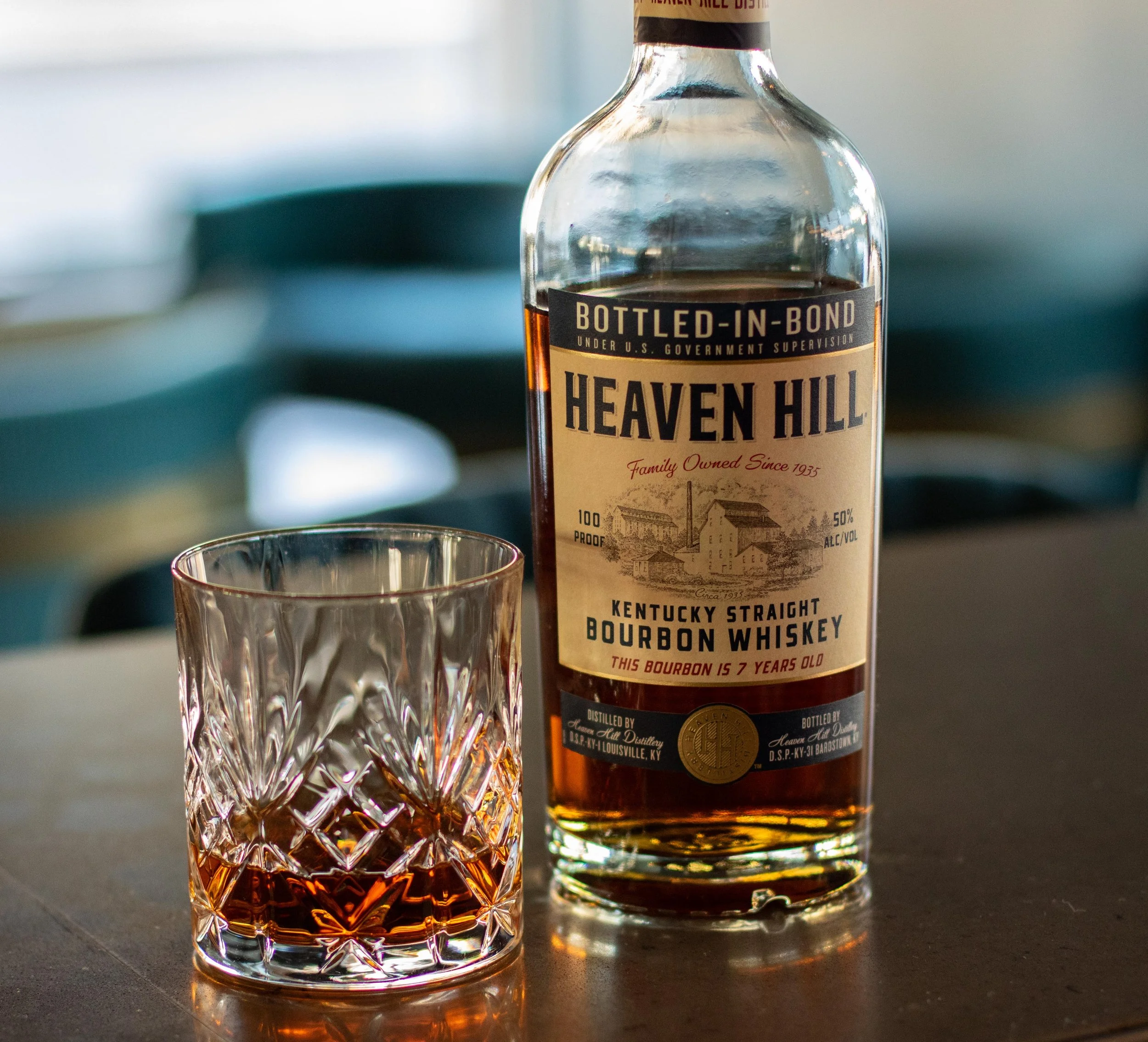 A bottle of Heaven Hill Kentucky straight bourbon whiskey and a glass with whiskey on a dark surface, with a blurred background.