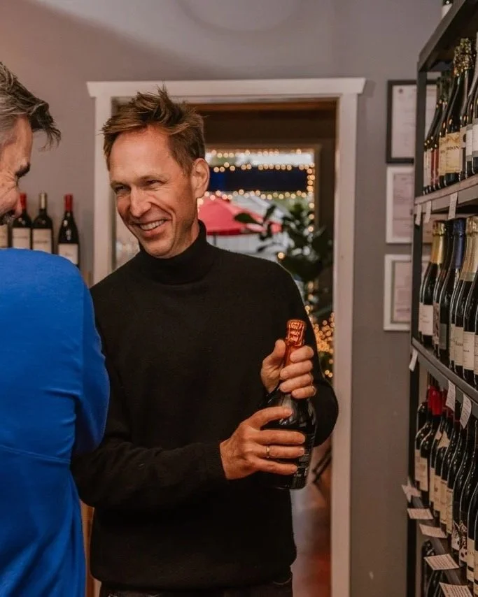 A man smiling while holding a bottle of wine or champagne in a wine shop, with shelves of bottles on his right and another person partially visible on the left.
