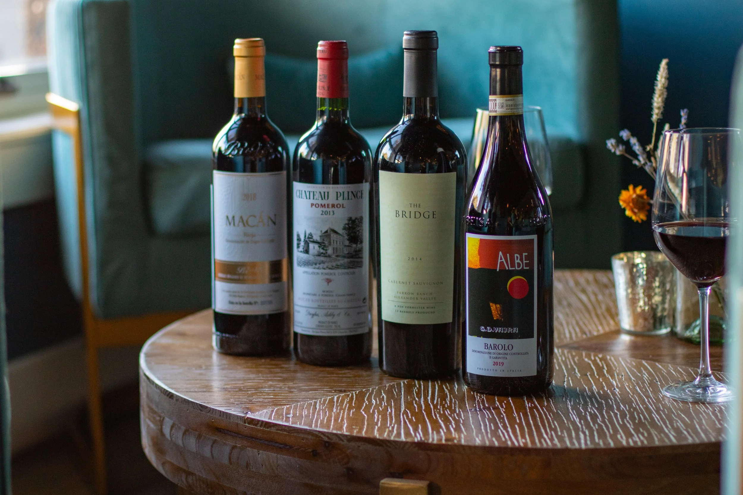 Four bottles of red wine placed on a wooden table, with a glass of red wine and small vases with flowers nearby.