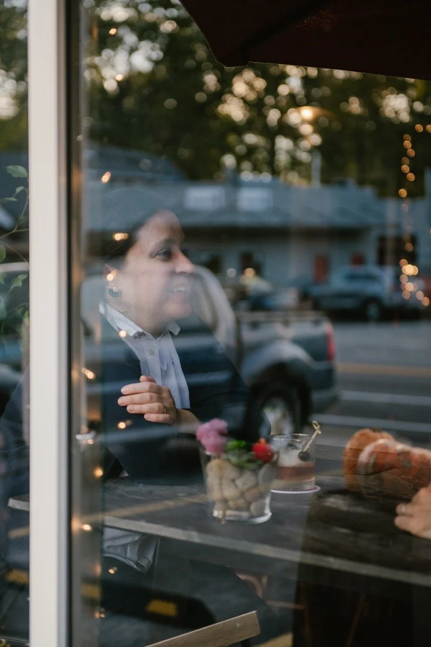 A woman in a black blazer and white shirt sitting at a table inside a cafe, seen through a glass window, with outdoor scene and cars in the background.