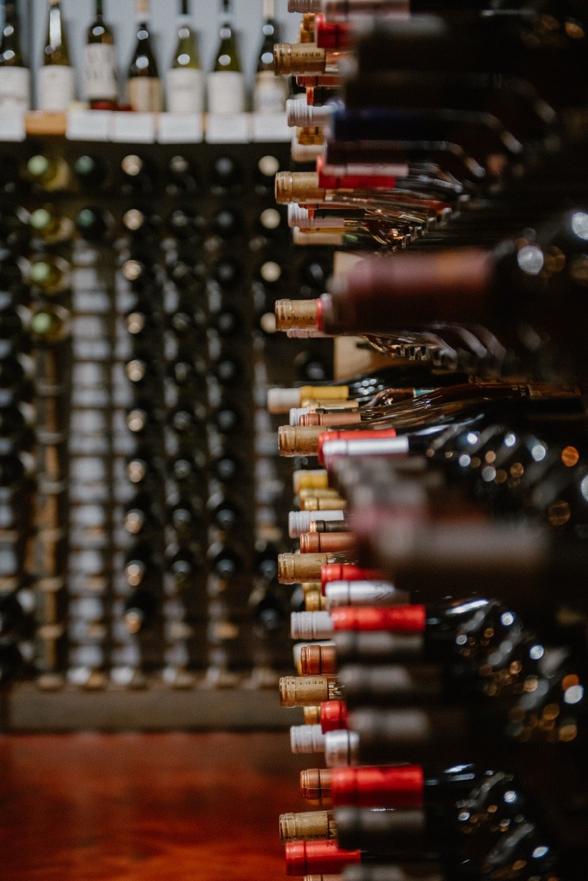 A wine cellar with bottles of wine stored on horizontal racks, with the focus on bottles with various colored caps on the right side, and a blurred background of additional bottles on the left.