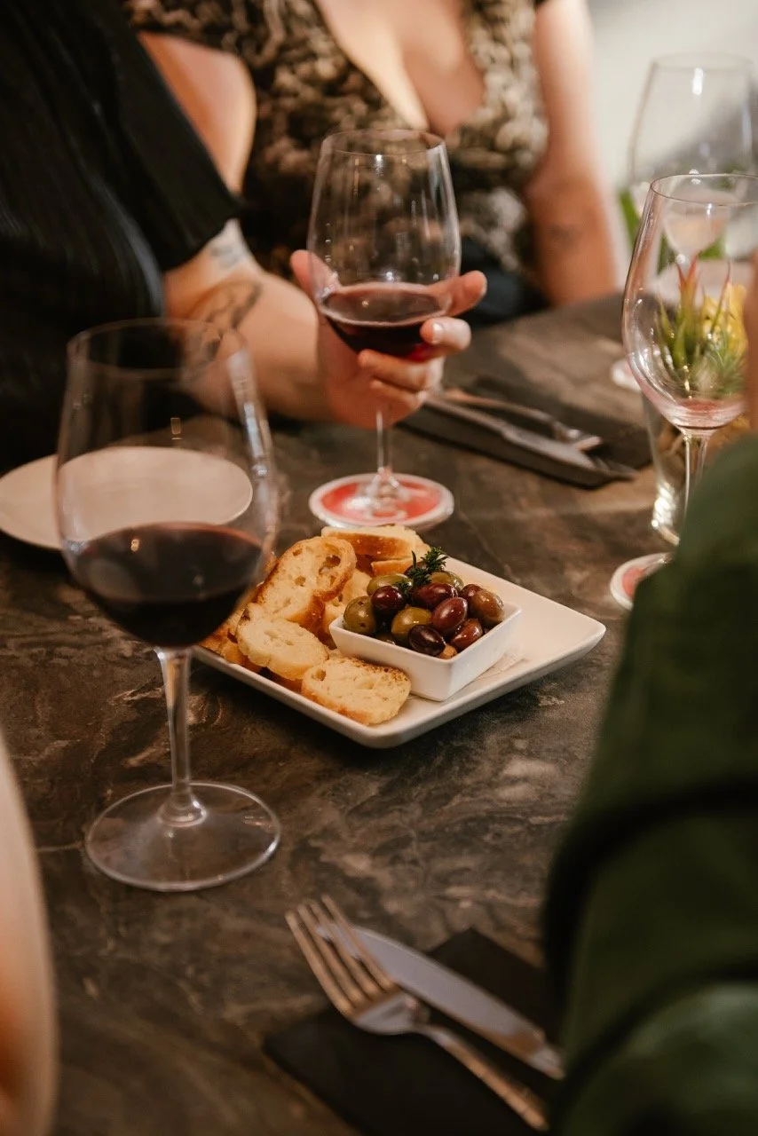 A person holding a glass of red wine, with a plate of bread and olives on a dark table surrounded by other glasses in a dining setting.
