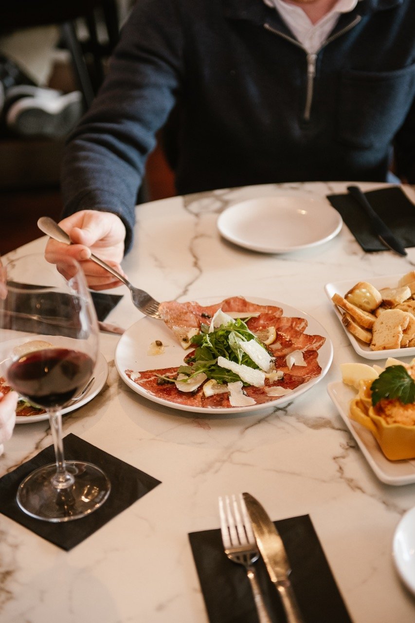 Person using fork to pick up cured meat and greens on a plate, with glass of red wine and other dishes on a marble table.