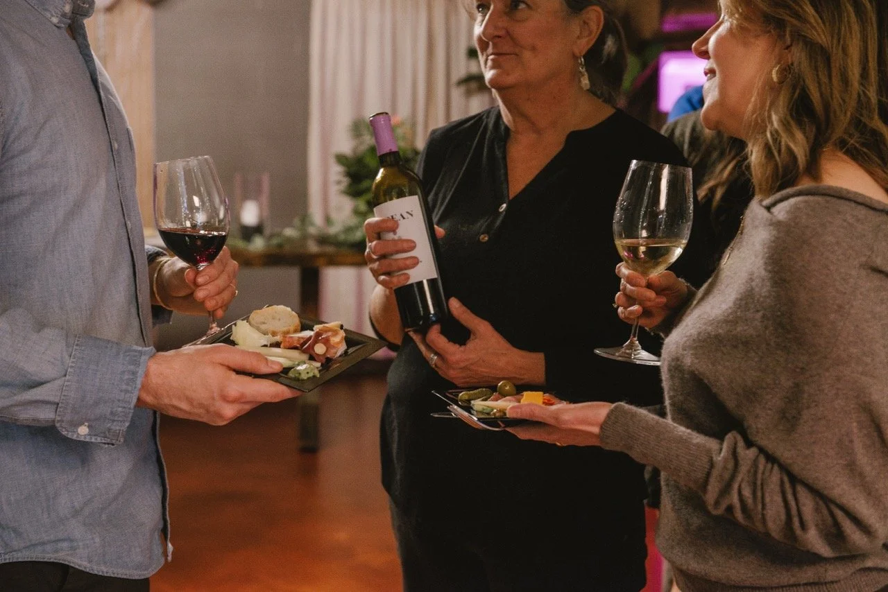 Three people at a social gathering holding wine glasses and plates of cheese, crackers, and snacks, engaged in conversation.