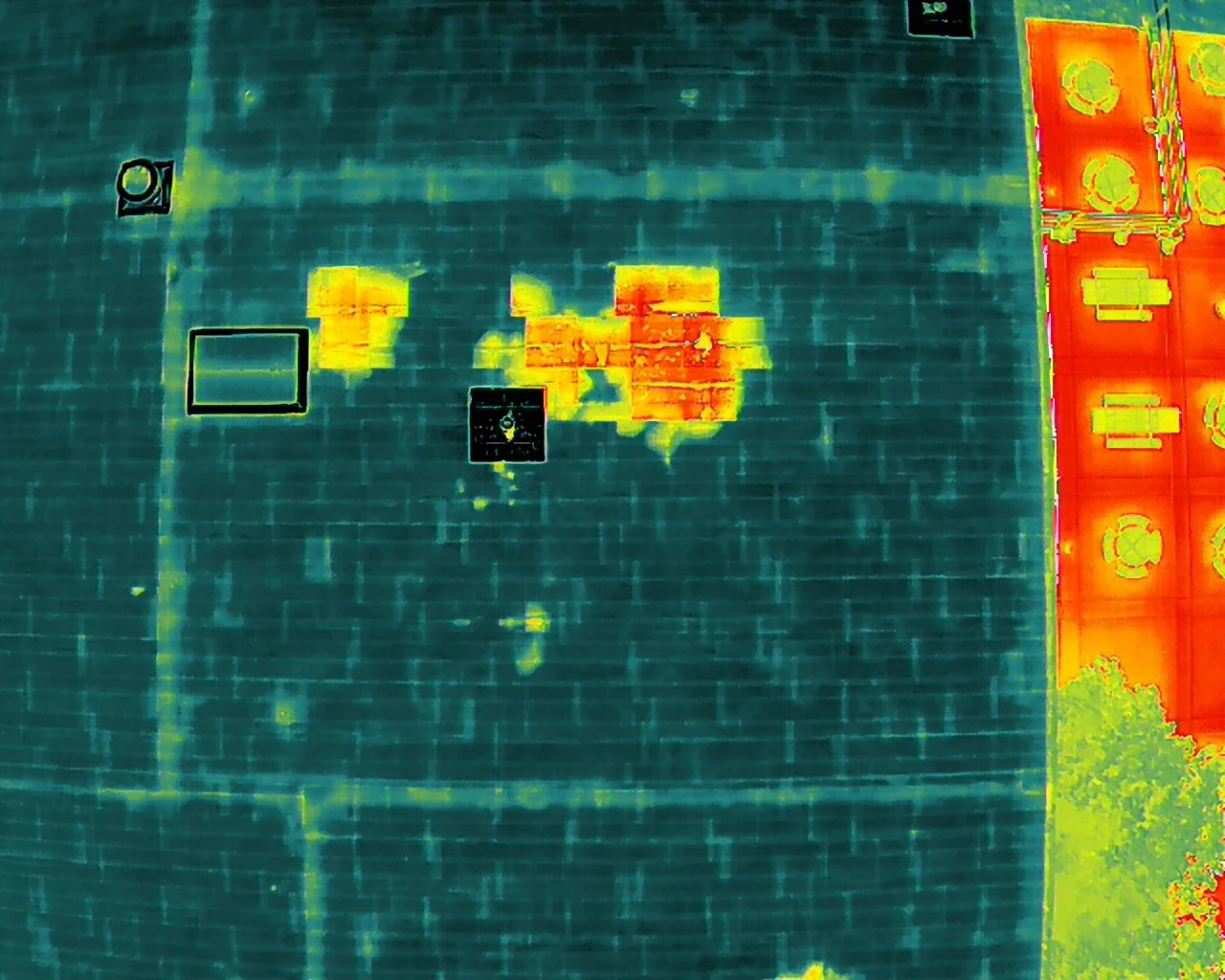 Infrared thermal image of a dark-colored rooftop with several warm spots and black outlined areas, next to a brightly lit area with various objects or structures on it.