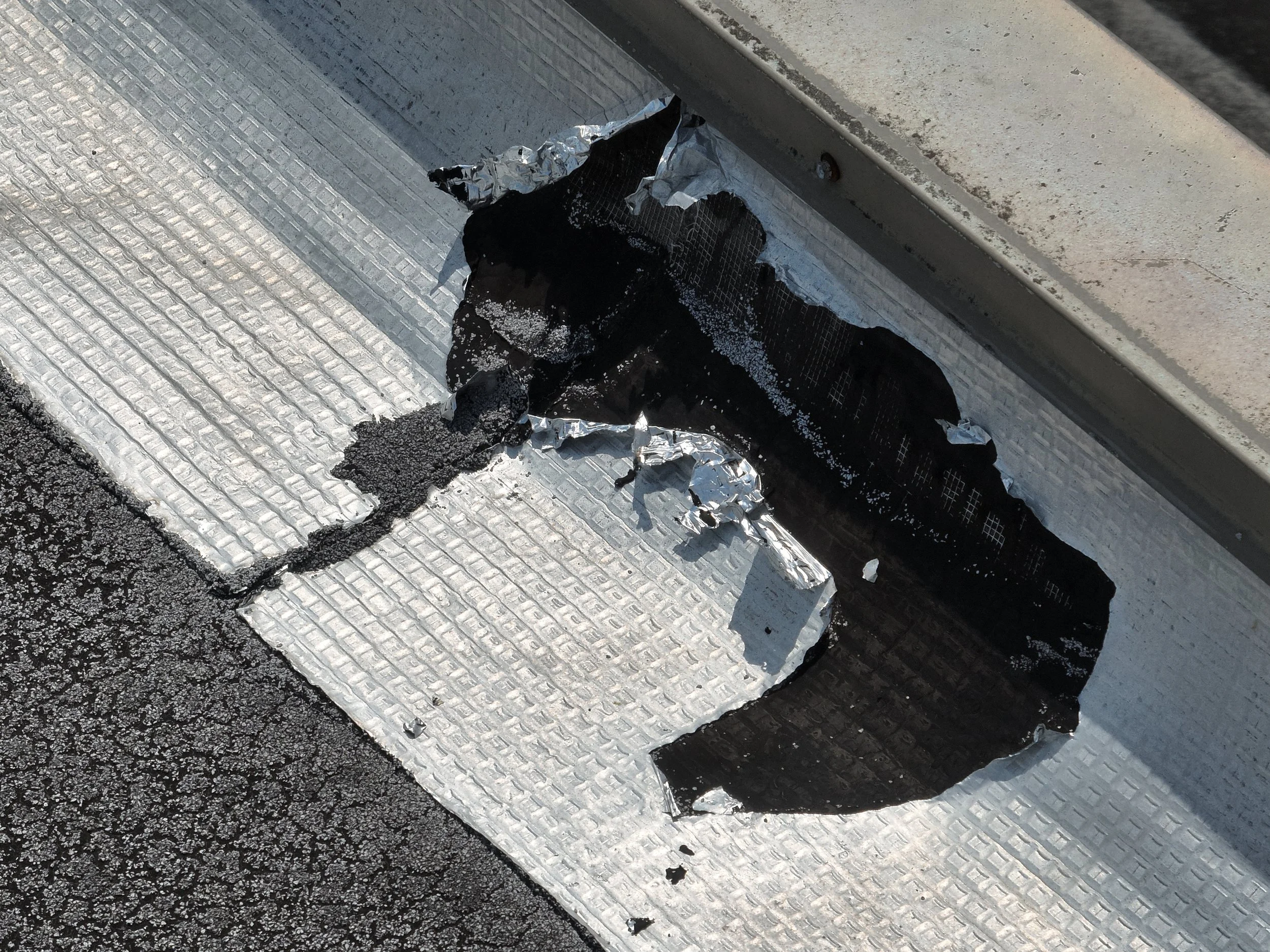 Close-up of damaged roofing material, including torn aluminum foil and black tar-like substance, on a textured rooftop surface with a gravel edge.