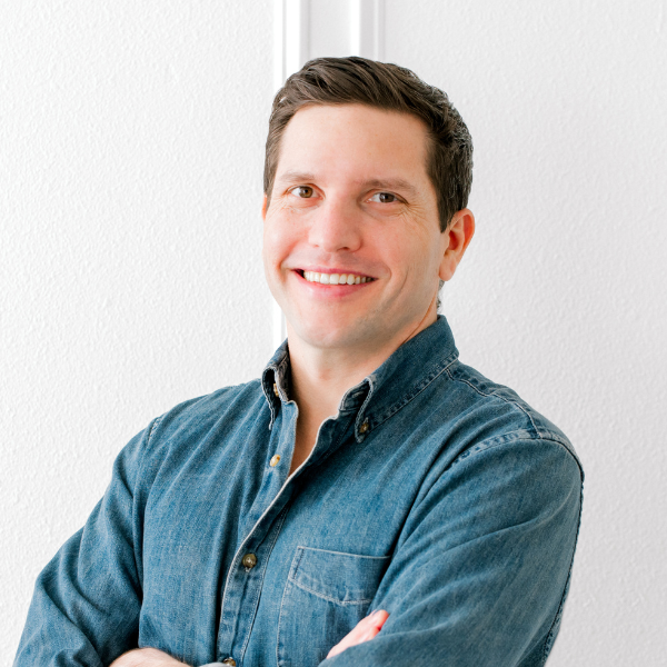 Smiling man with short brown hair wearing a denim shirt, standing against a plain white wall.