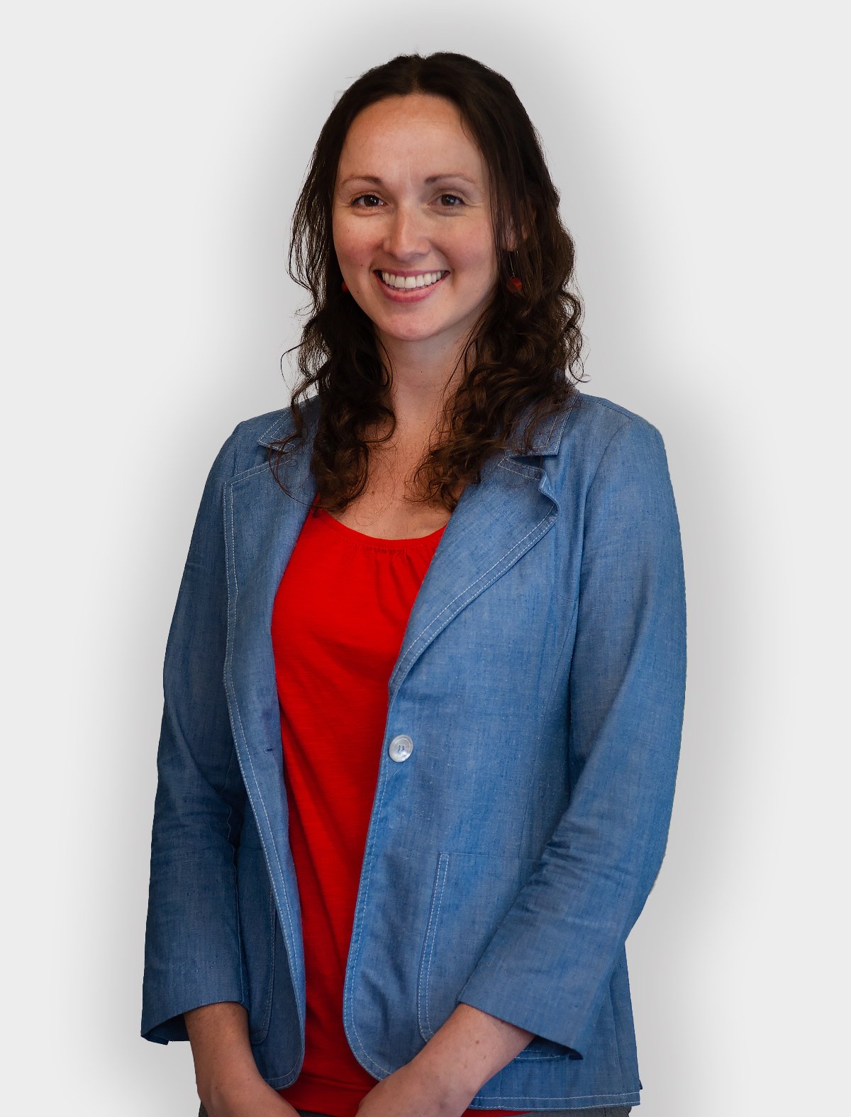 A woman with long curly brown hair, smiling, wearing a denim blazer over a red top, standing against a plain light gray background.