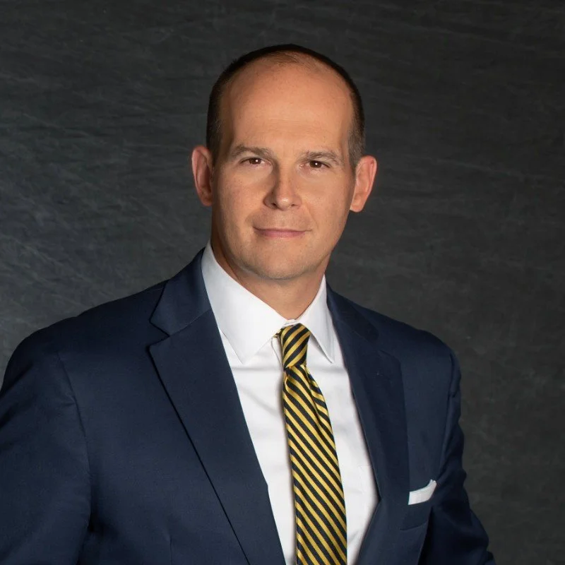 Professional headshot of a man in a dark suit, white shirt, and striped tie, against a dark textured background.