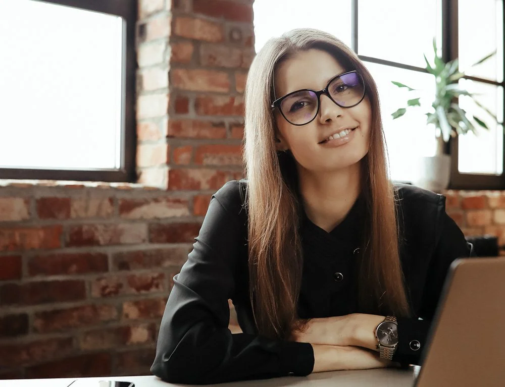 Woman with glasses smiling at desk in office with brick wall background