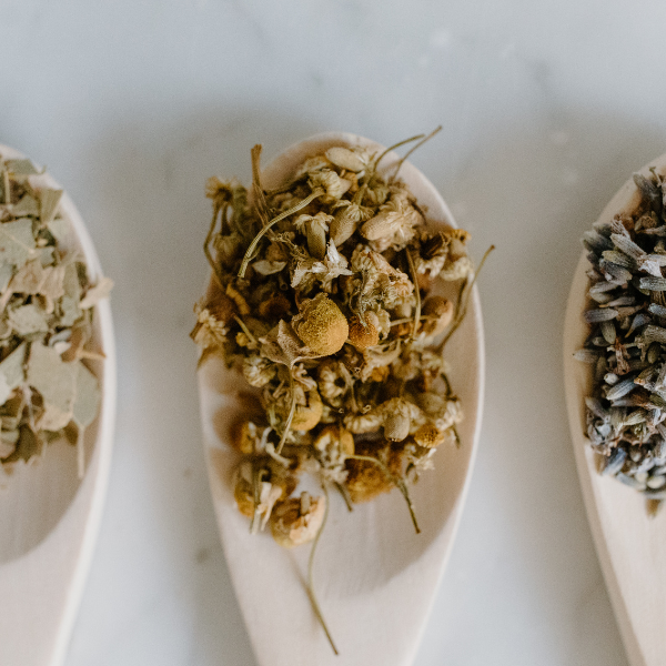 Dried herbal flowers on a white ceramic spoon, with other dried herbs on a similar spoon in the background.