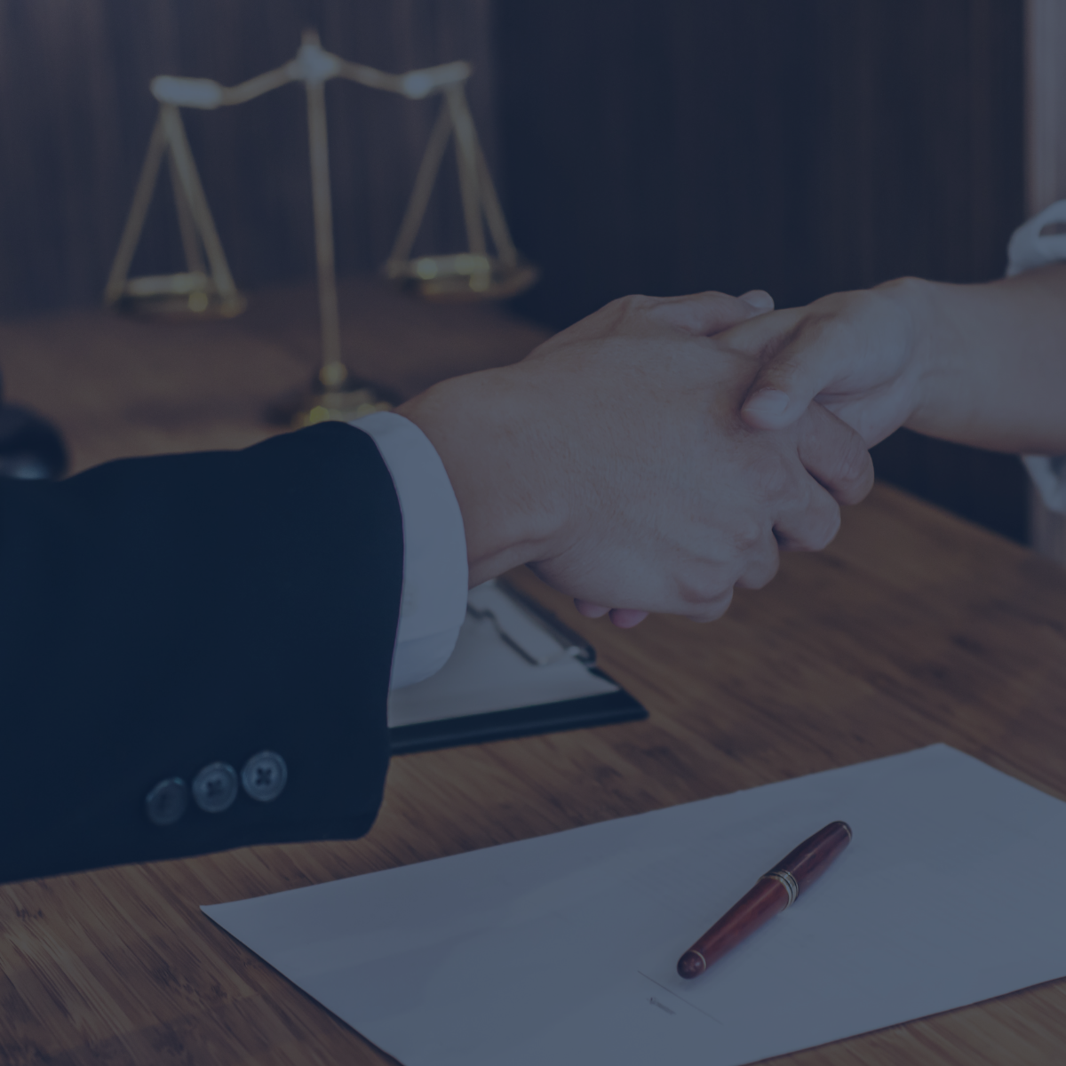 Zwei Personen besiegeln mit einem Handschlag eine Vereinbarung. Im Vordergrund liegen ein unterschriftsreifes Blatt Papier mit Füllfederhalter, im Hintergrund steht eine Justitia-Waage.