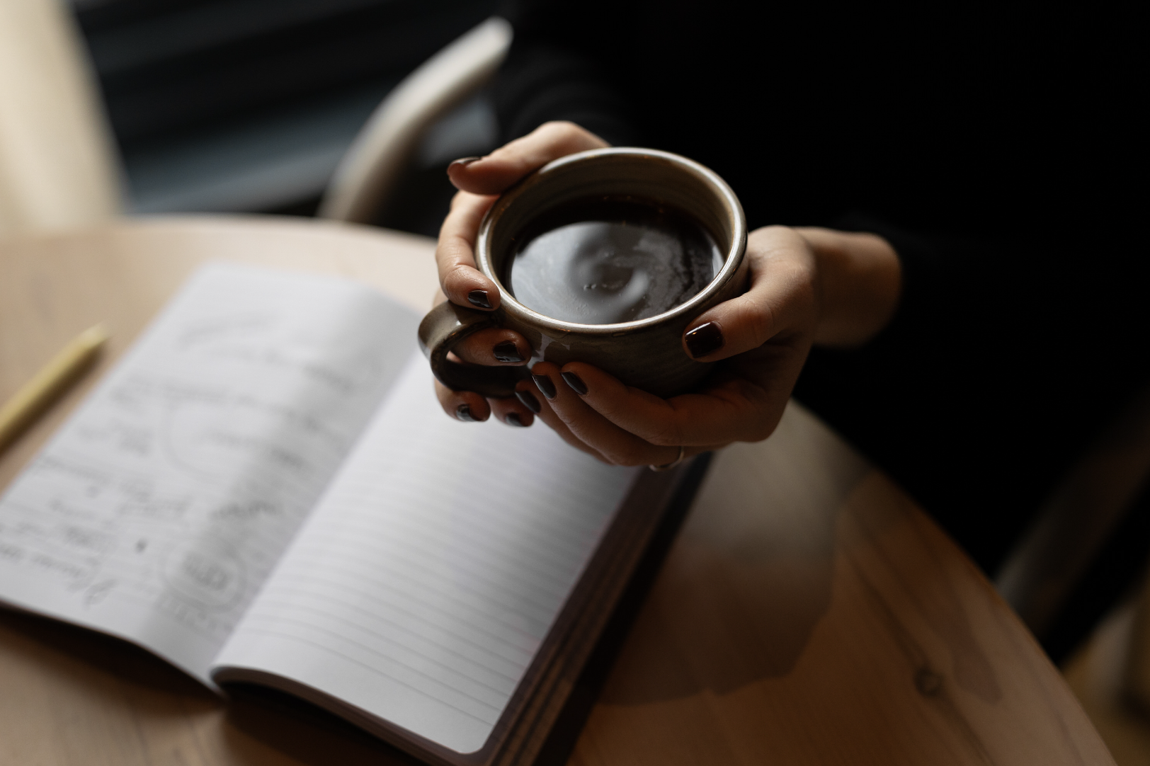 A person holding a ceramic mug filled with a hot beverage over an open notebook on a wooden table.