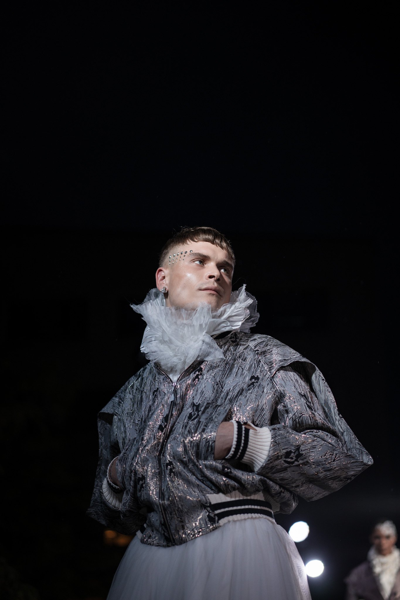 Fashion model wearing a metallic jacket with a high ruffled collar on a dimly lit runway show.