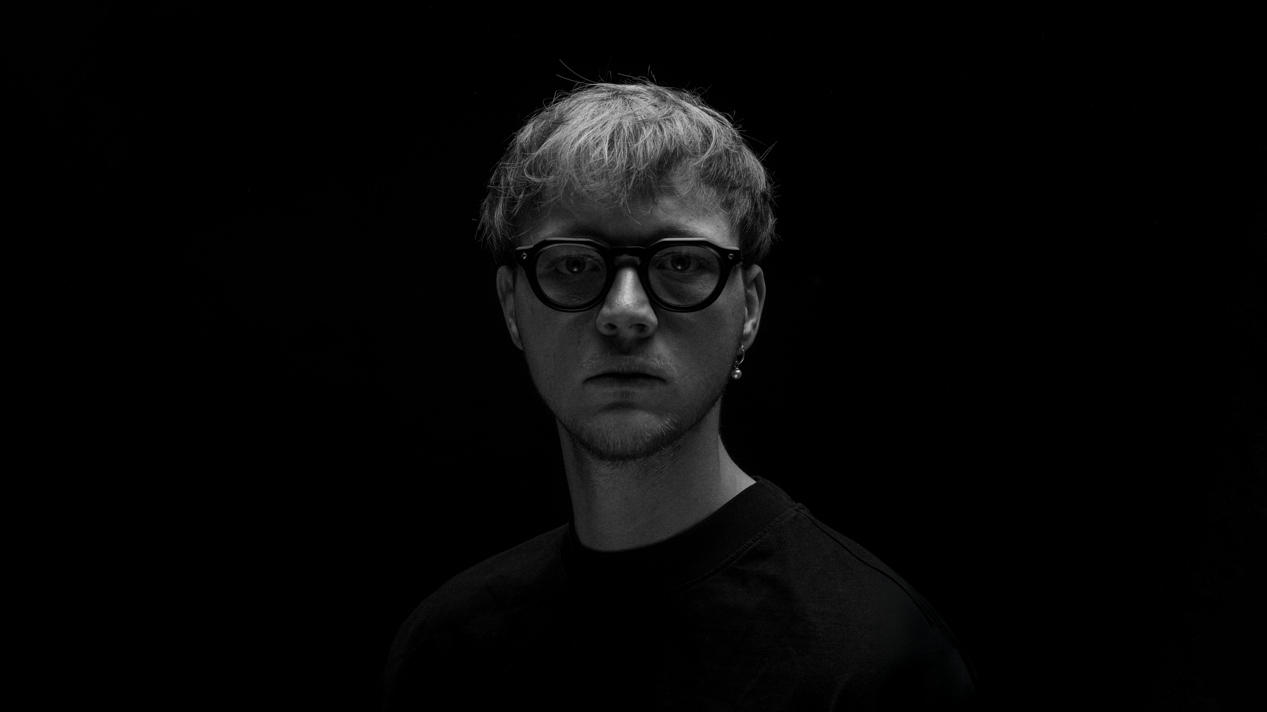 Black and white portrait of a young man with glasses, short hair, and a serious expression, against a dark background.