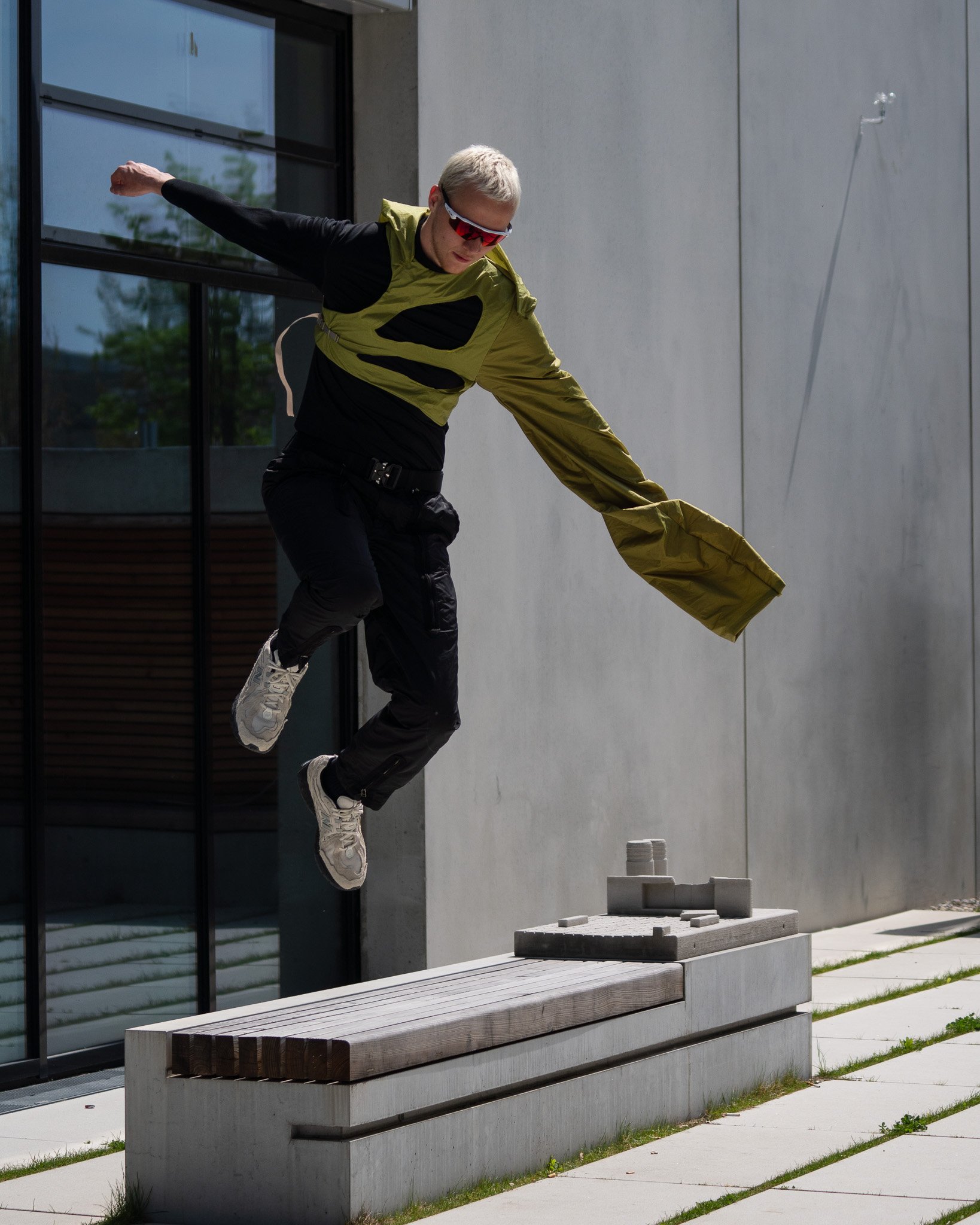 Person wearing a green and black outfit jumping mid-air next to a concrete bench, outdoors.