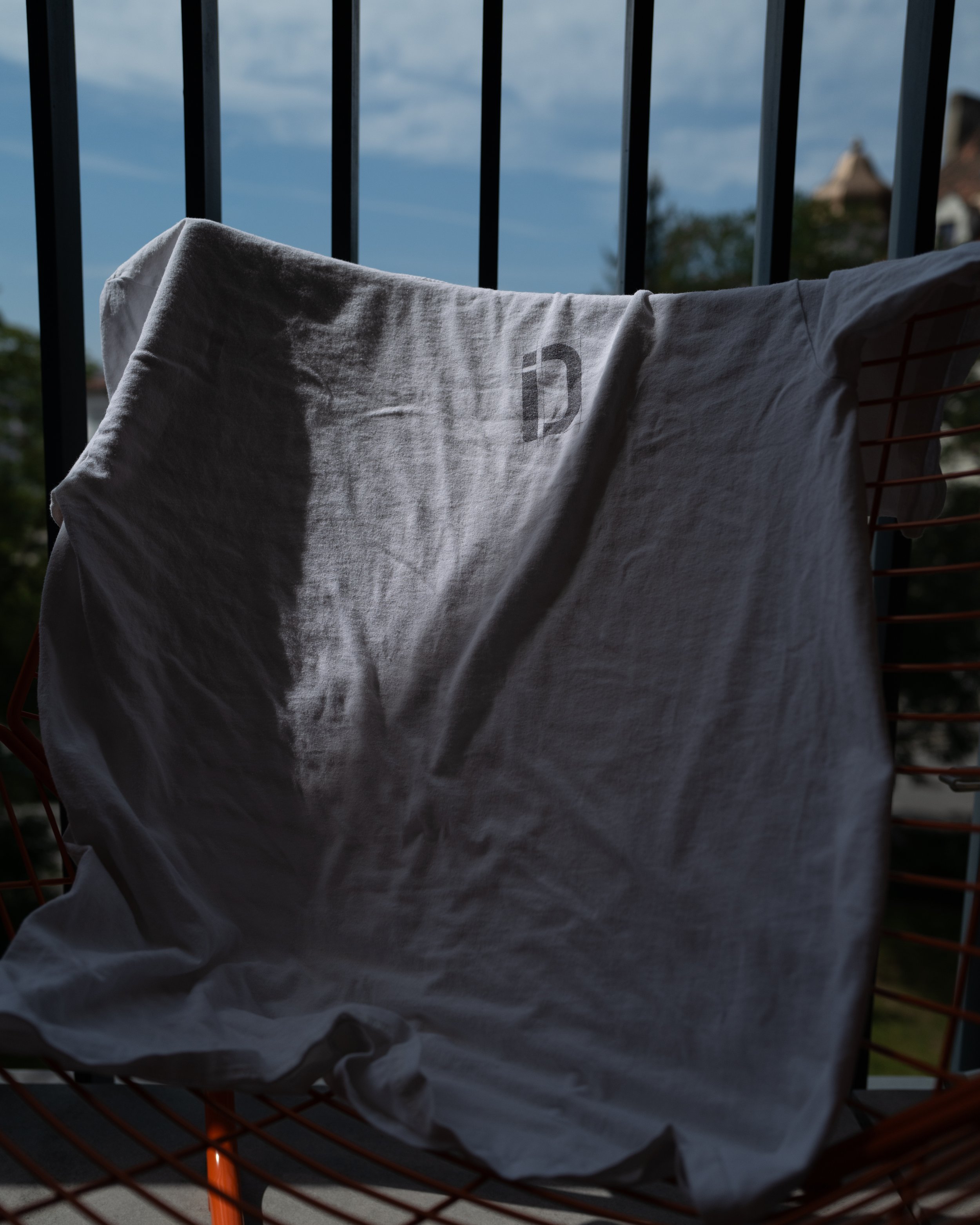 White t-shirt draped over a wire chair on a balcony, with metal railings and an outdoor view in the background.