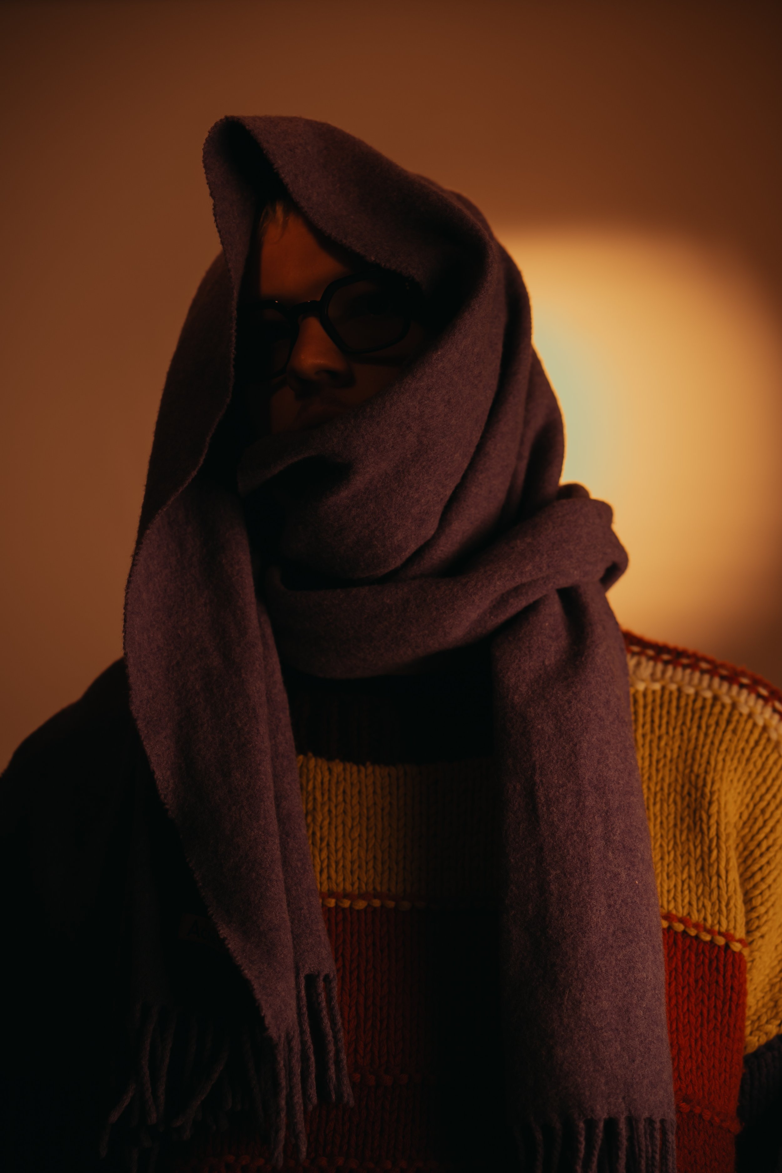 Person wearing glasses wrapped in a thick scarf with a colorful knit sweater, dim lighting, and warm tones.
