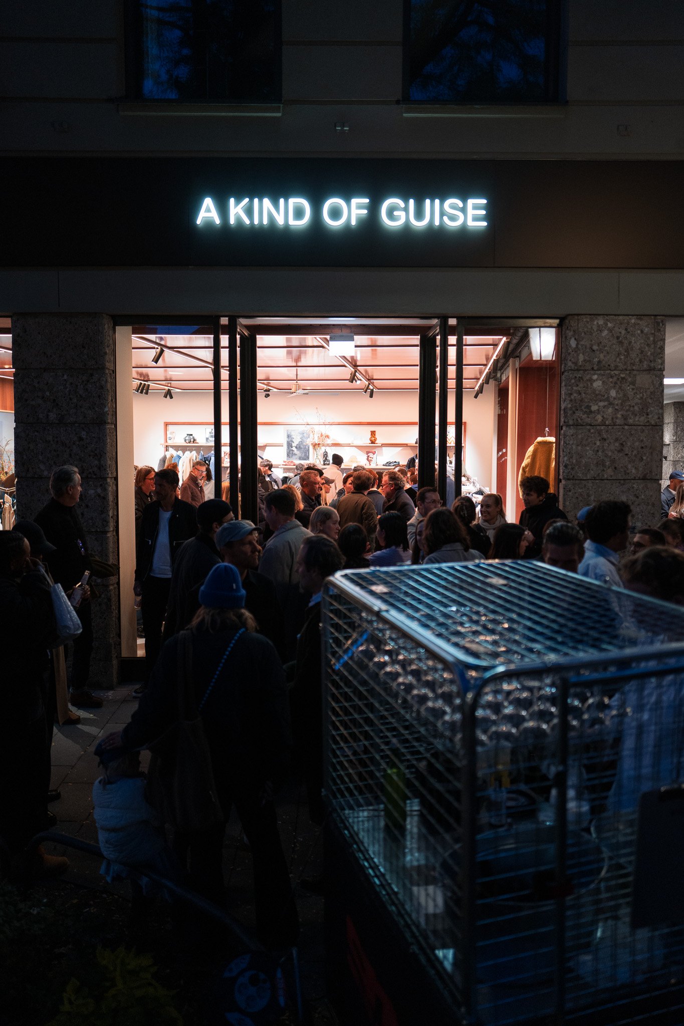 Crowd gathered outside a store with "A KIND OF GUISE" sign above entrance, evening setting, visible lighting inside the store, glass rack in foreground.