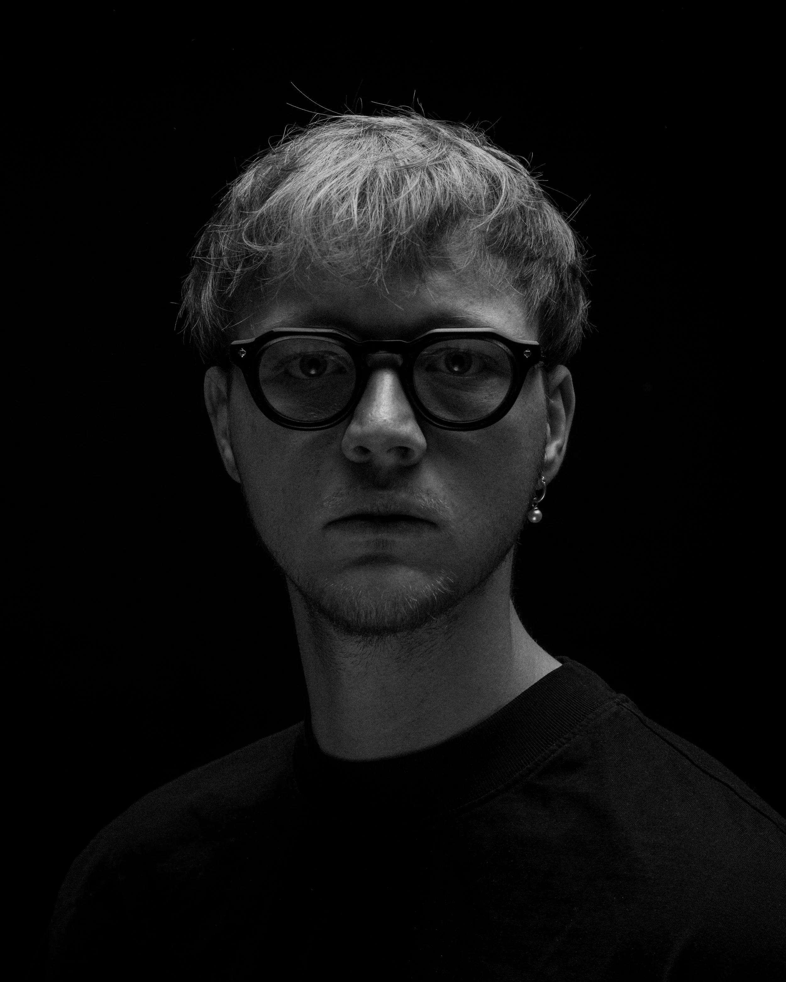 Black and white portrait of a young man with glasses, earring, and short hair, looking directly at the camera against a dark background.