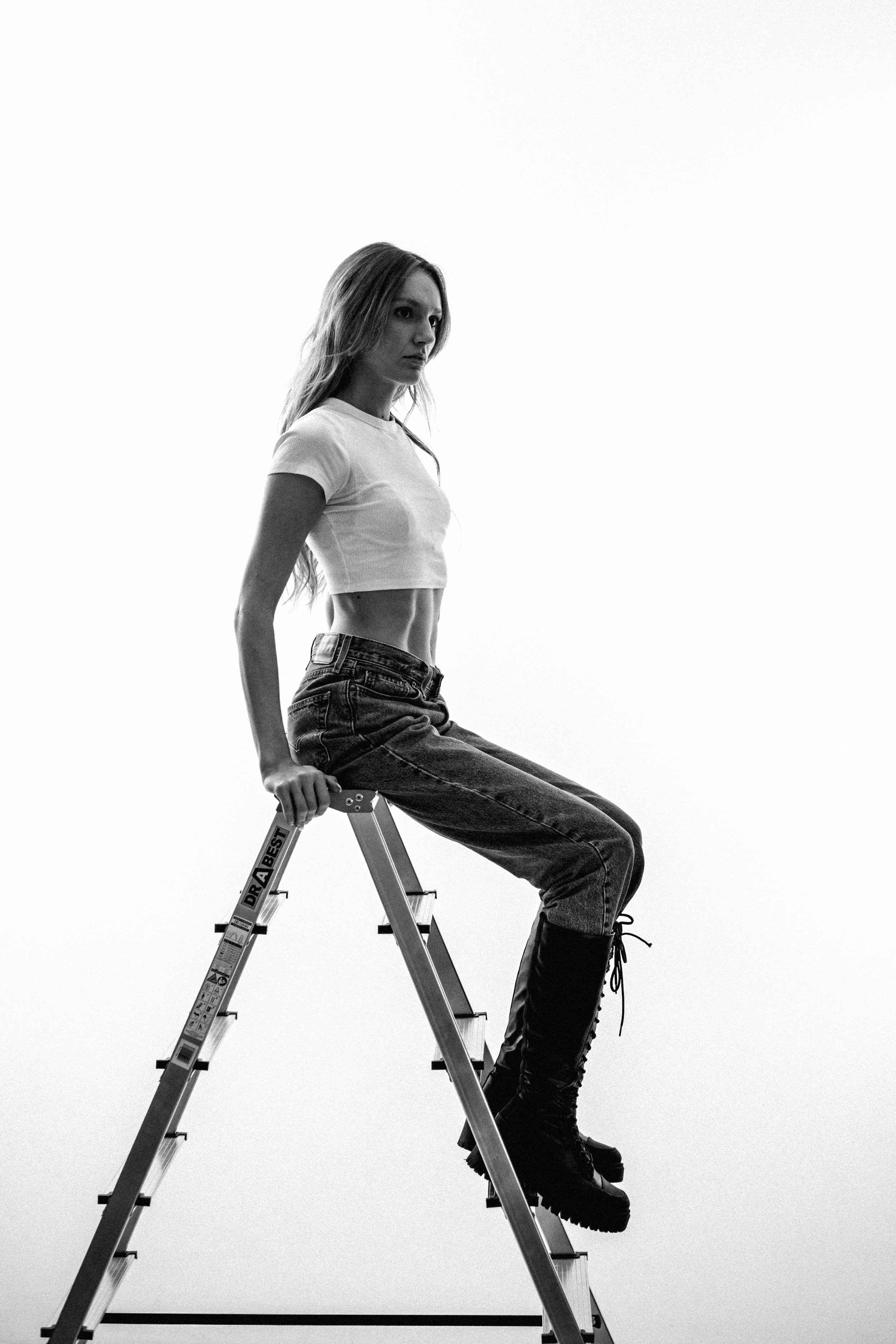 Black and white photo of a woman sitting on a ladder, wearing a white crop top, jeans, and boots.
