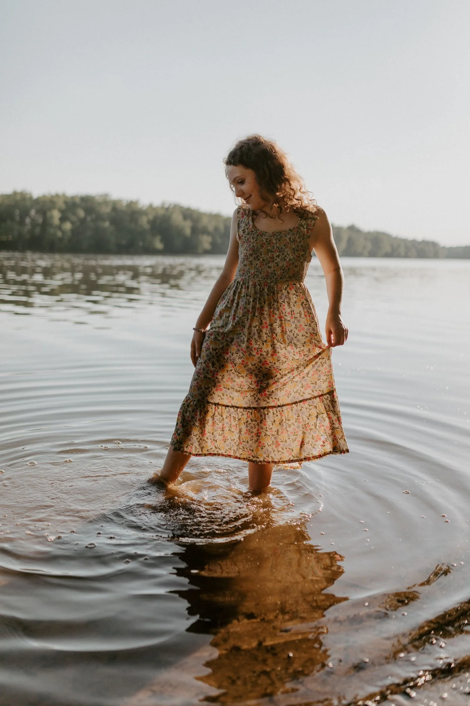 Natural, unposed photo of a little girl in a dress standing in a river at sunset, capturing an organic, storytelling moment in soft golden light