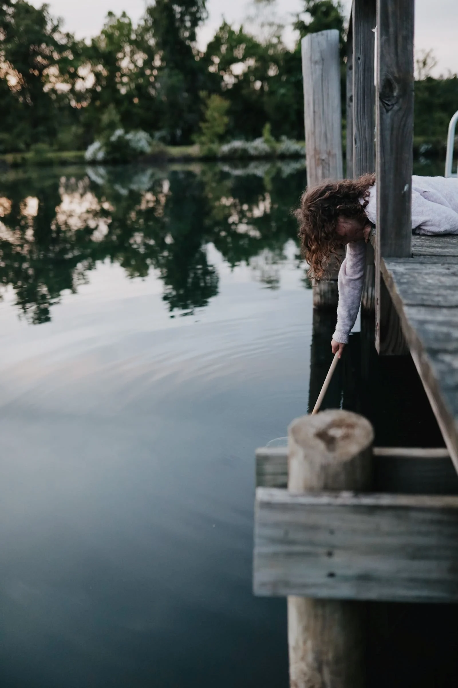 curly haired girl leaning over a dock with a net trying to catch turtles in a pond