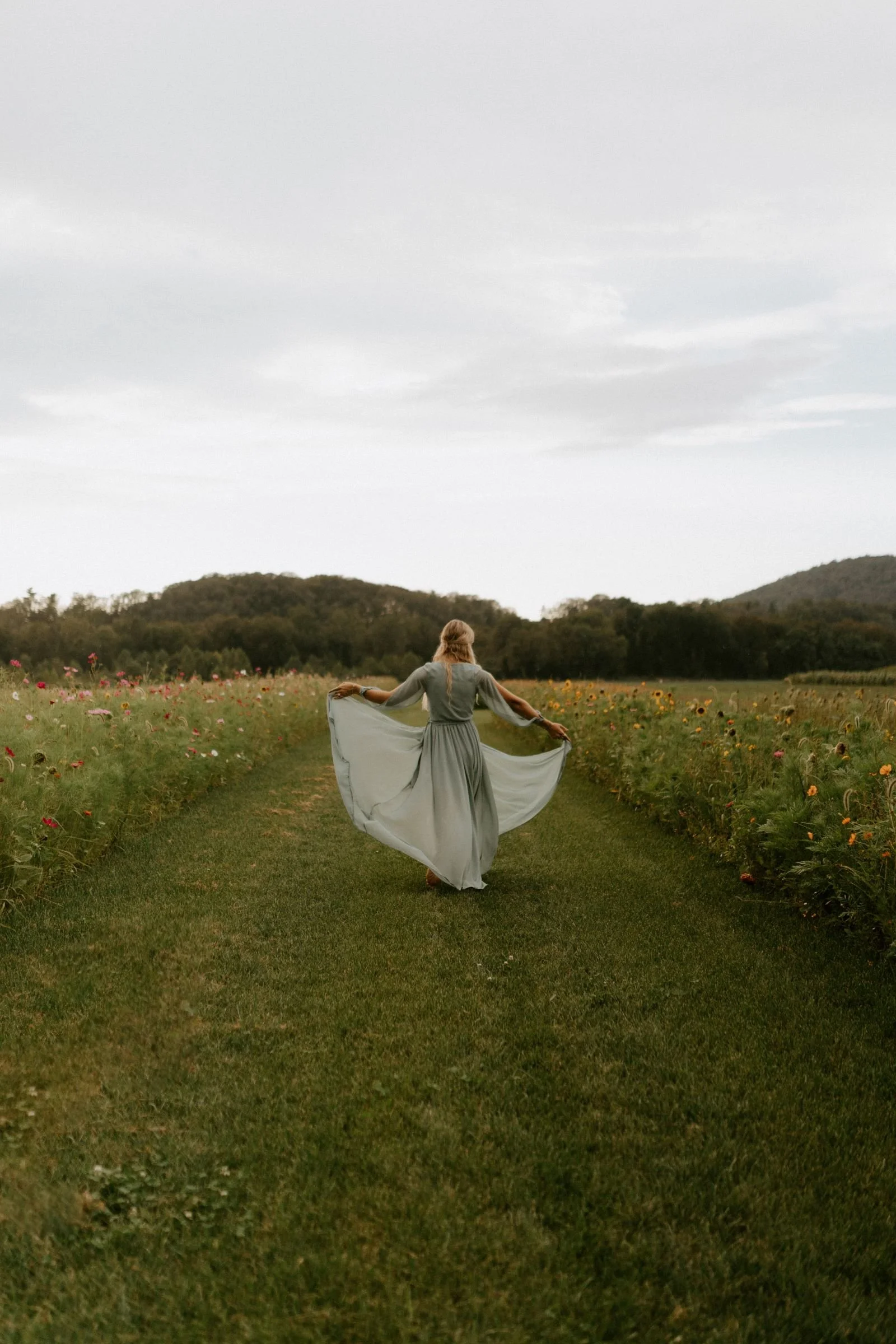 young woman holding out her light blue dress and dancing down a path flanked by flowers