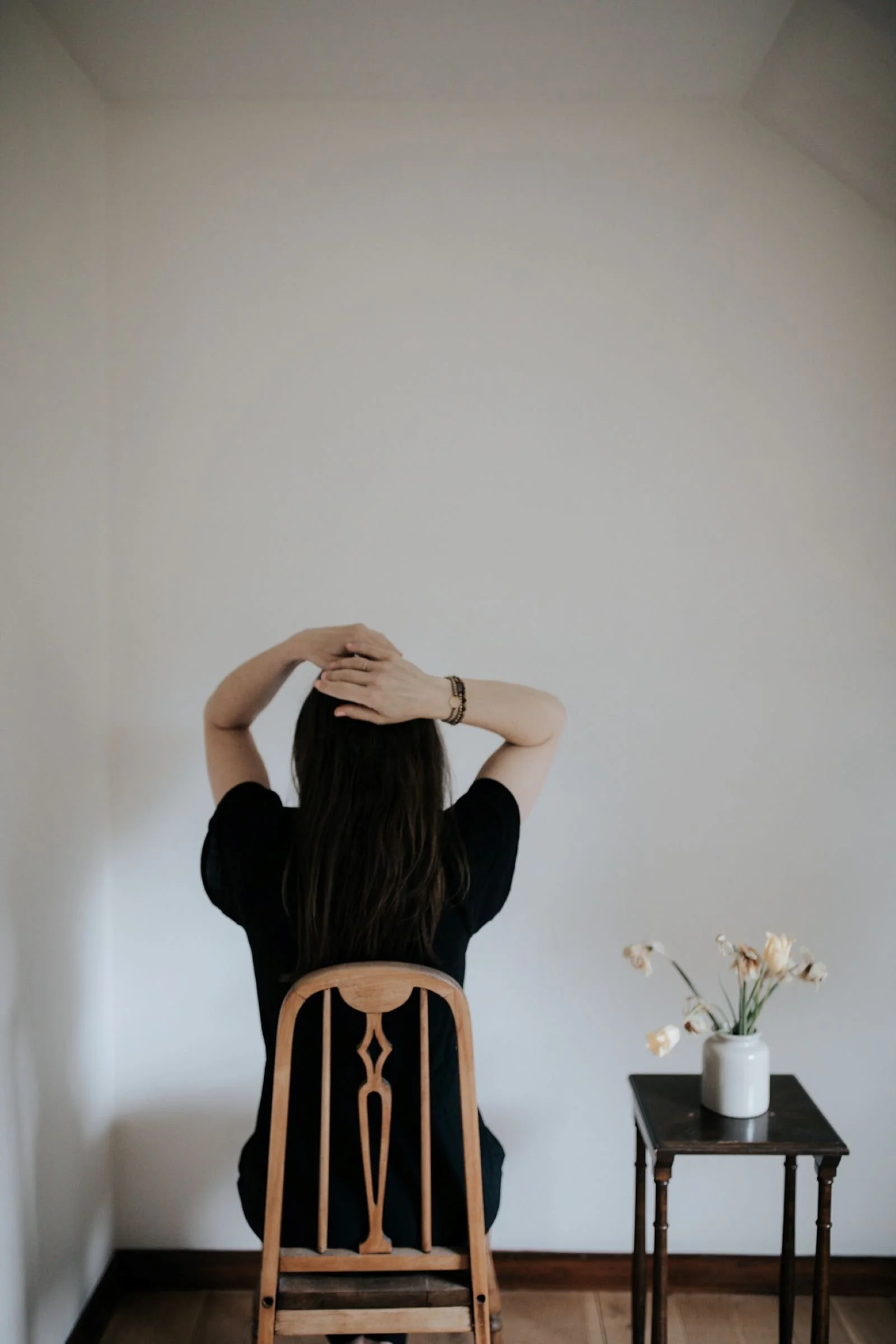 a woman sitting with her back to you in a chair and resting her hands on her head with a small table and white vase with flowers beside her
