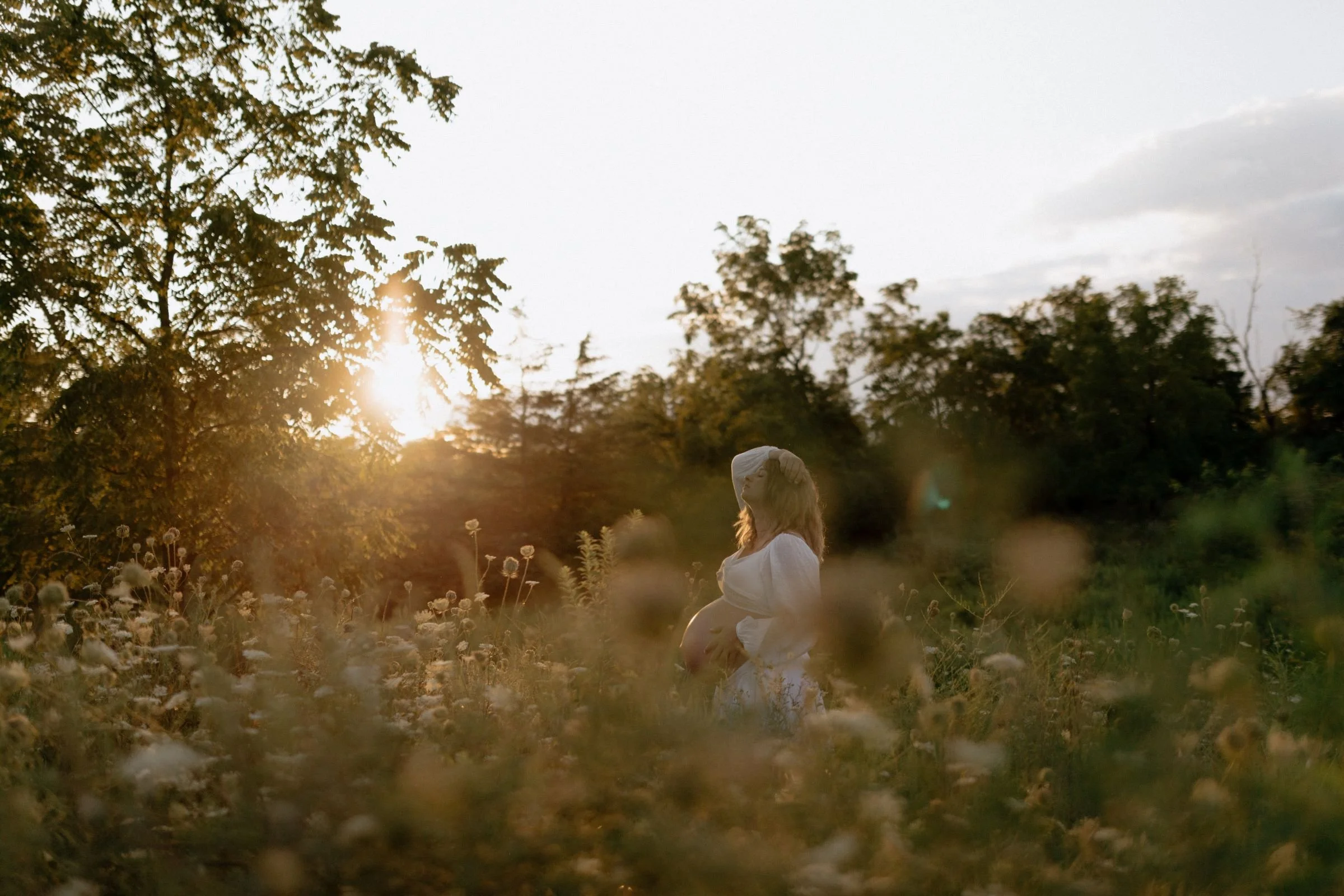During an unposed maternity session, a pregnant woman in a white dress stands in a flower field at sunset.