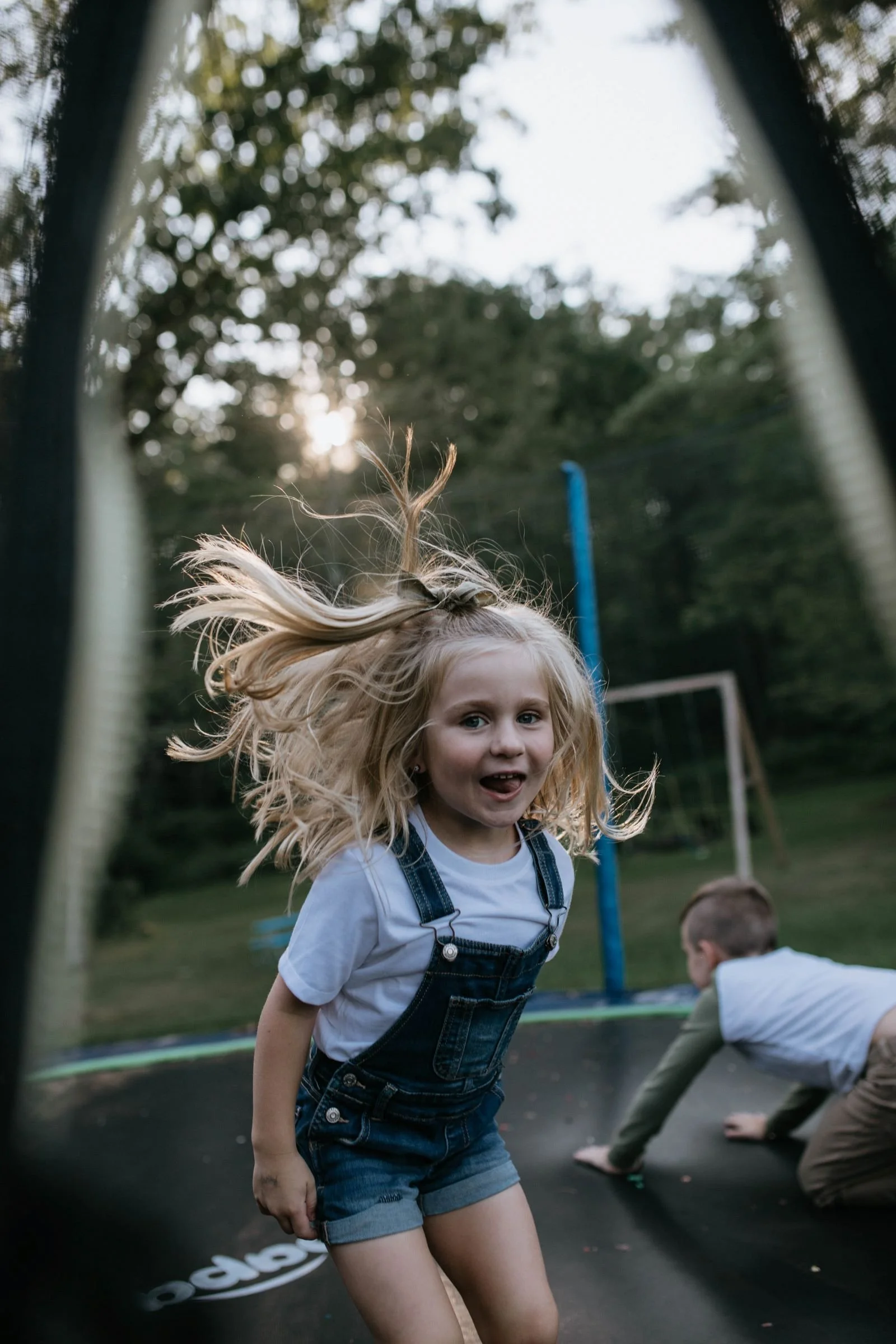 blonde five year old girl wearing overalls, jumping on a trampoline