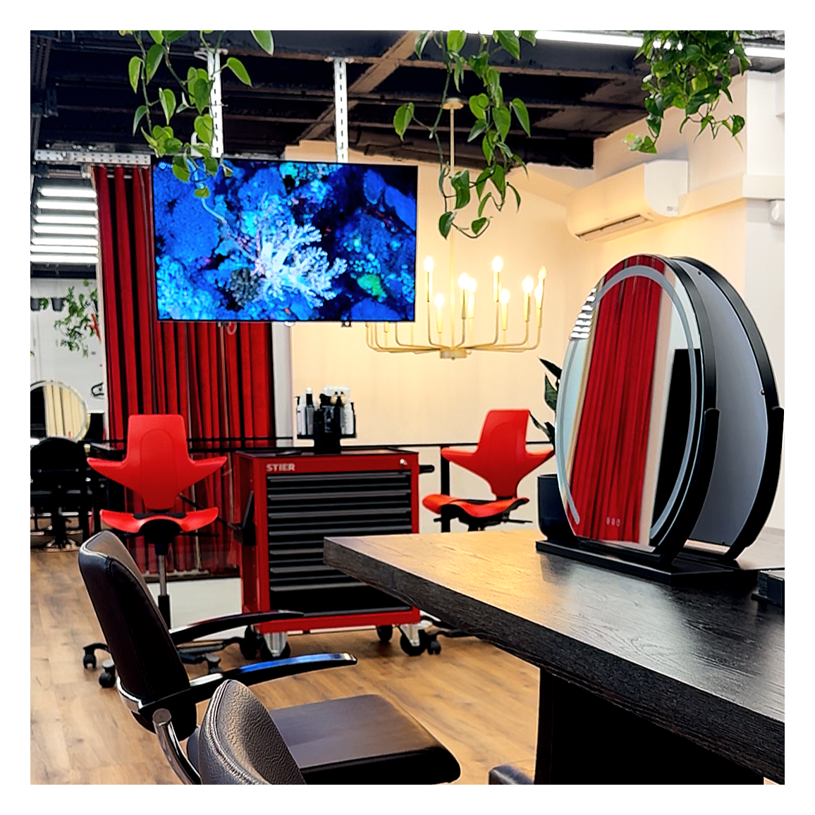 Ein modern eingerichteter Salon mit einem großen Fernseher, roten Stühlen und einem Spiegel auf einem schwarzen Tisch. Es gibt eine Deckenlampe und grüne Pflanzen als Dekor. Der Raum hat einen Holzfußboden und eine rote Vorhang. Ein Technik-Wagen ist sichtbar, während im Hintergrund die Decke offen ist.
