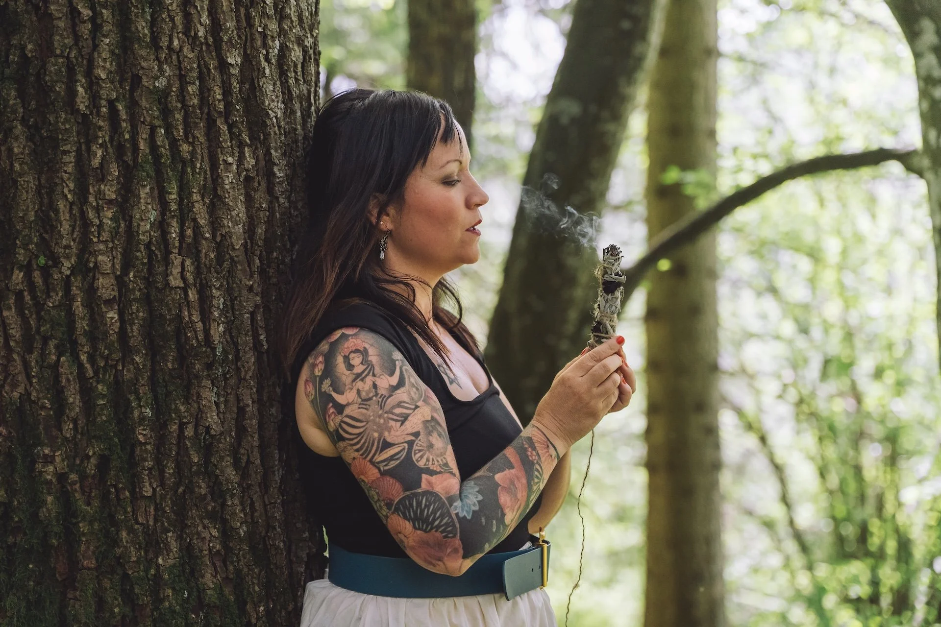 Akasha Chronik lesen lernen: Alina lehnt im Wald an einem Baum und räuchert mit einem Salbeibündel.