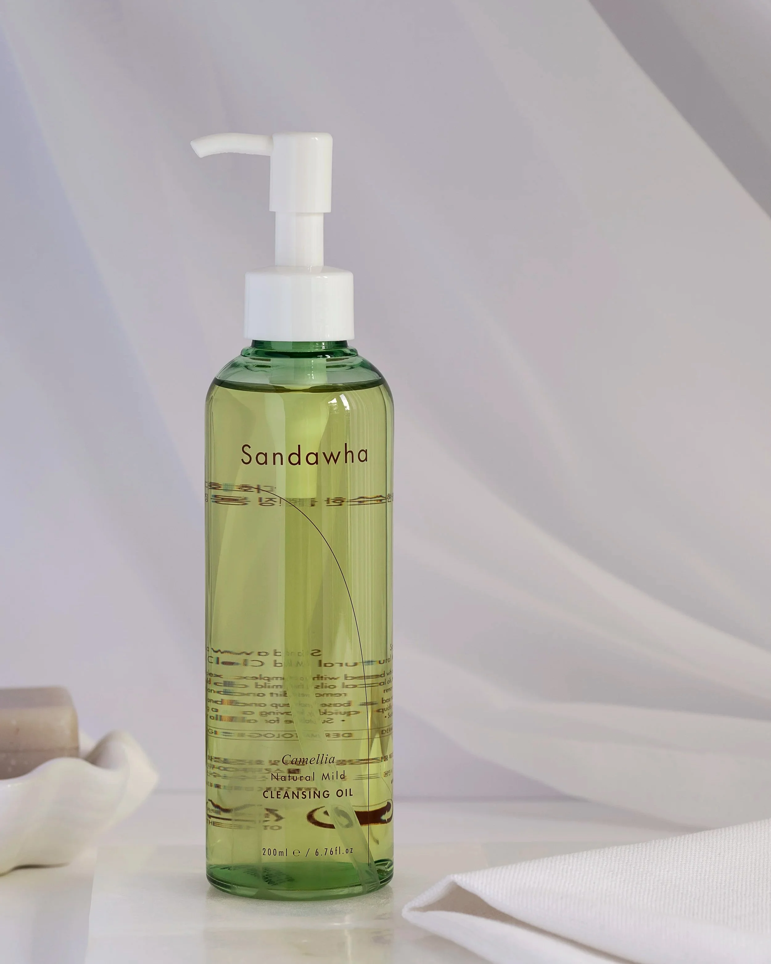 Green clear plastic bottle of Sandawha cleansing oil with a white pump dispenser on a white surface with a white cloth and seashells nearby.
