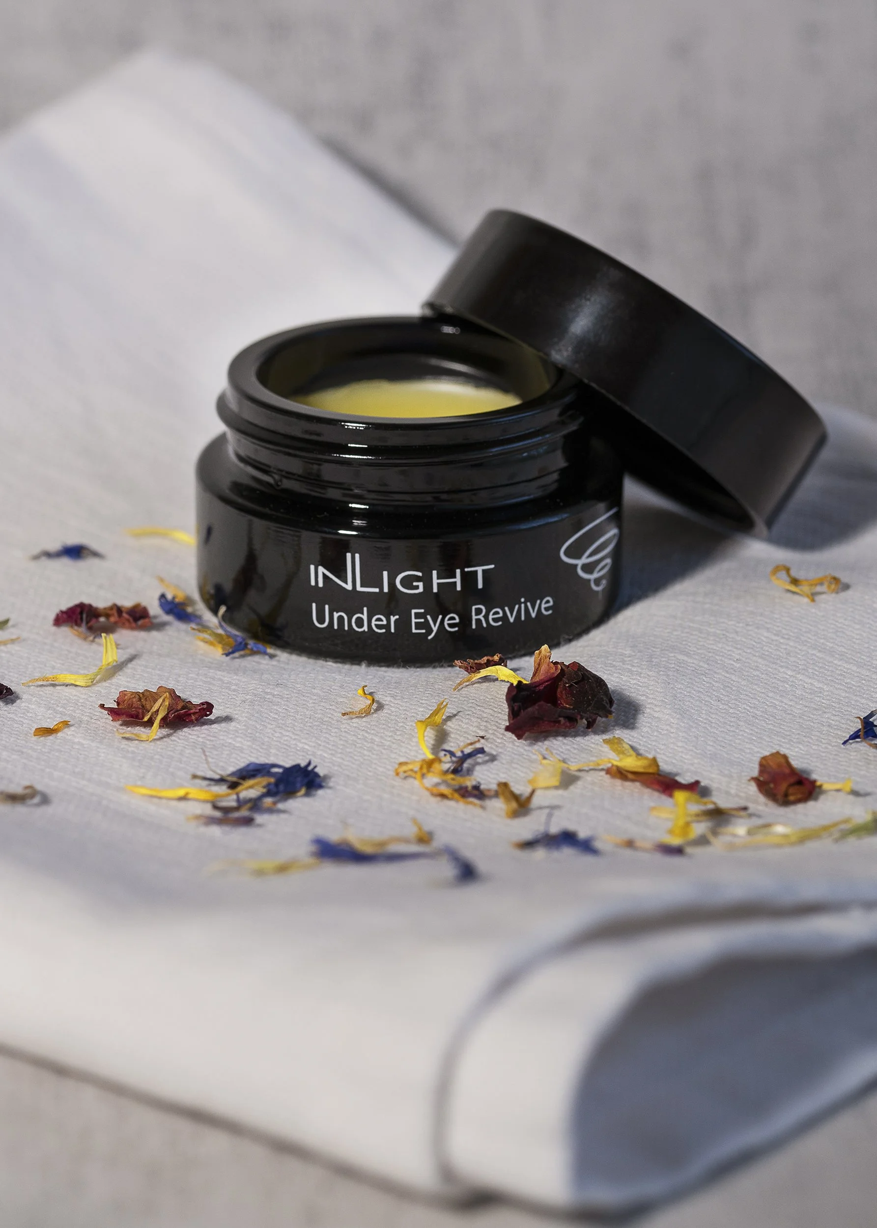 A black jar labeled 'INLIGHT Under Eye Revive' with a yellow cream inside, placed on a white cloth with scattered dried flower petals.