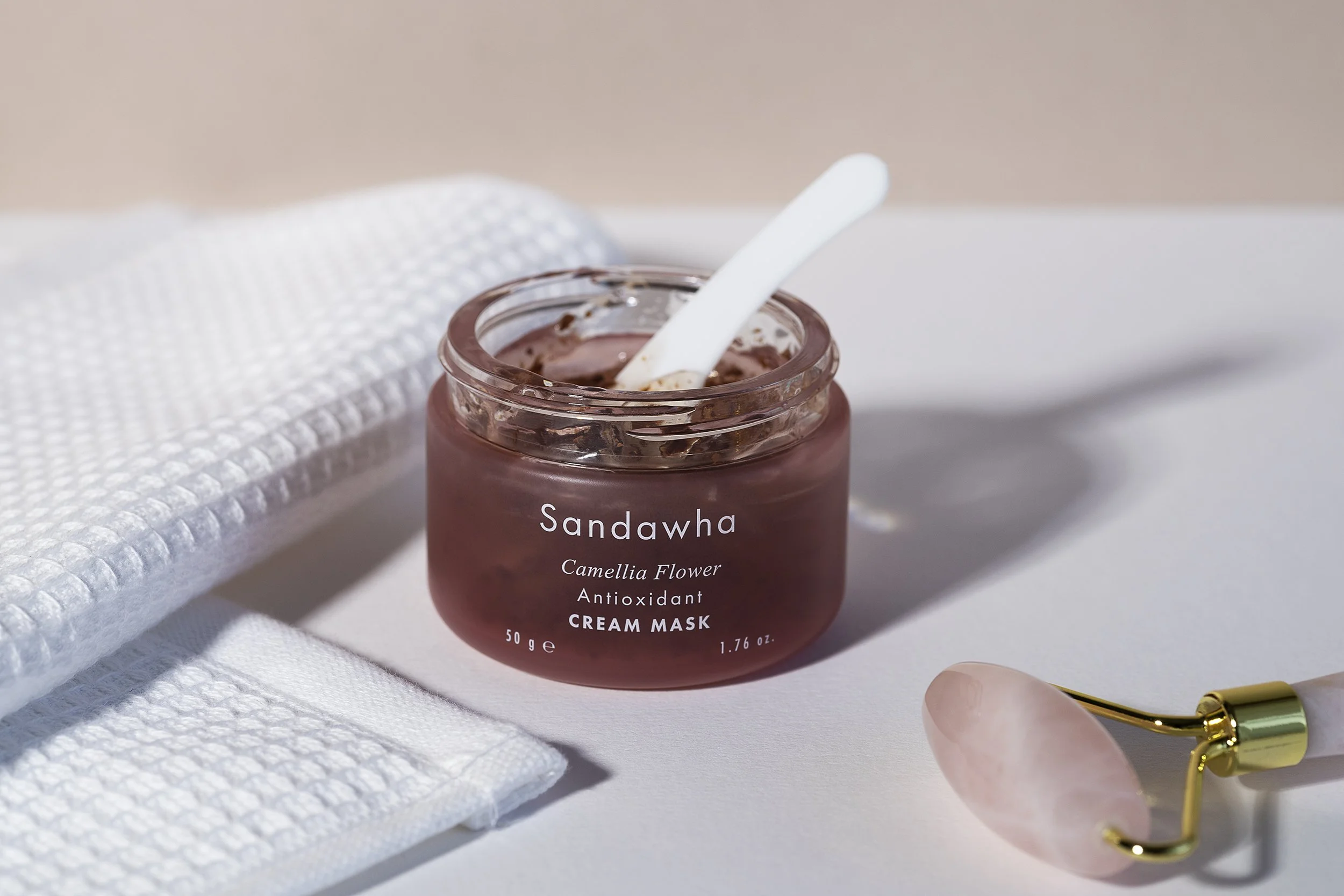 A jar of Sandawha Camellia Flower antioxidant cream mask with a spatula inside it, placed on a white surface next to a rose quartz facial roller, a white textured cloth, and a white towel.