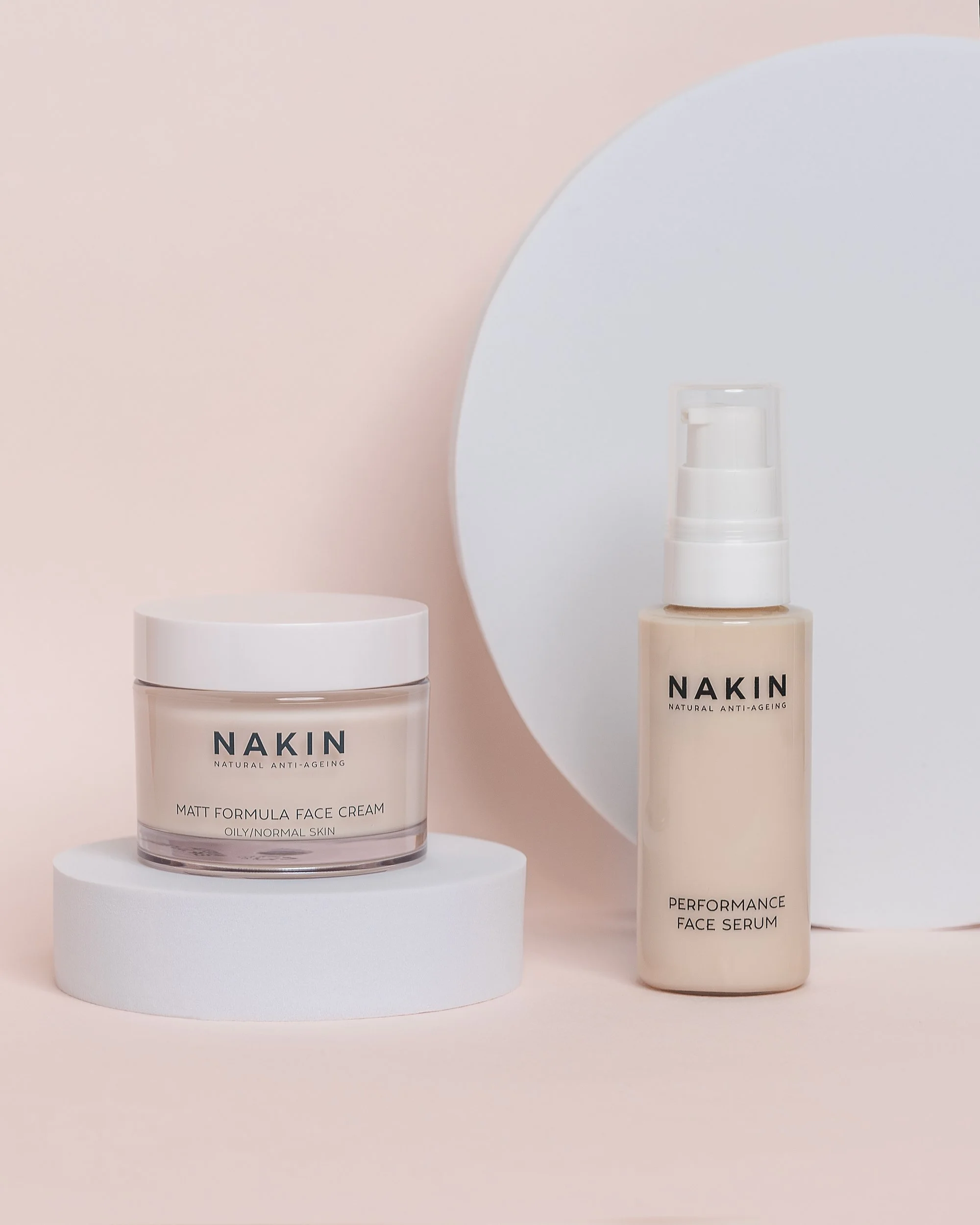 NAKIN face cream and face serum bottles on white cylindrical platforms against a neutral pink background.