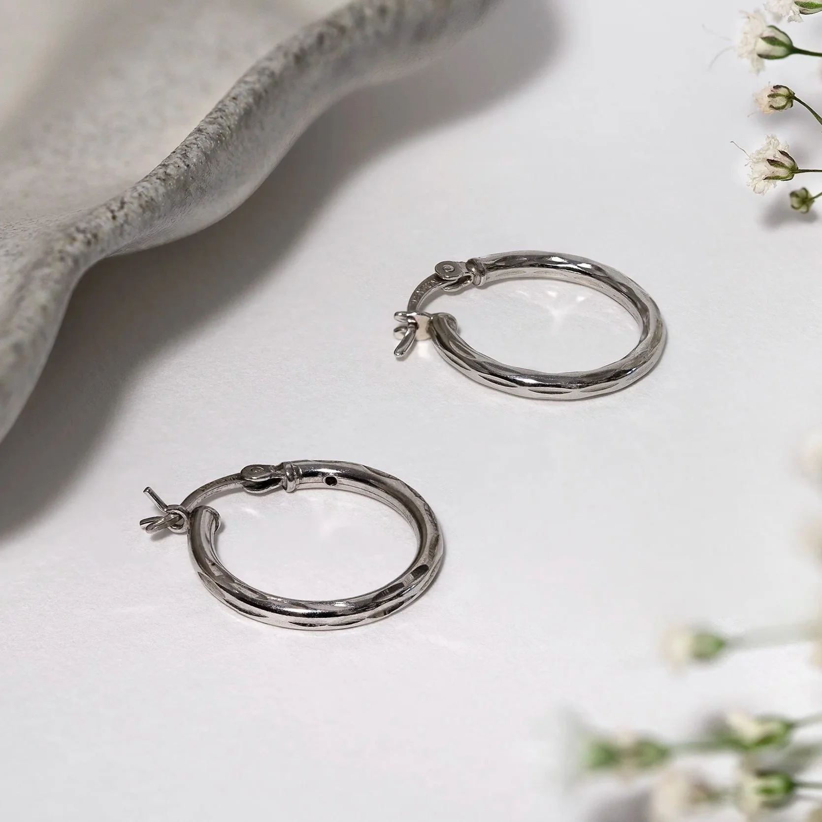 A pair of silver hoop earrings placed on a white surface with a textured gray object and small white flowers in the background.