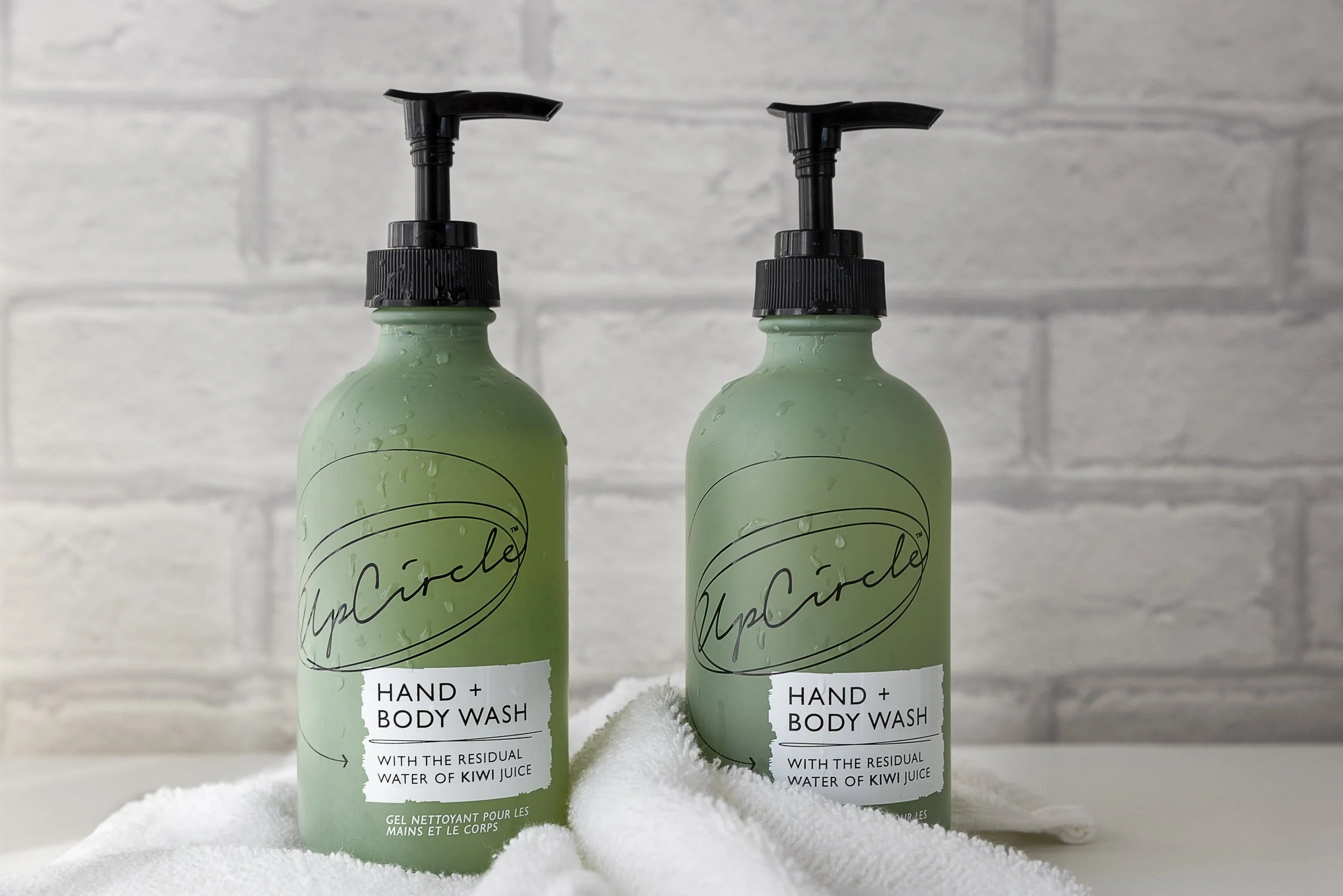 Two green bottles of YeapCicle hand and body wash with black pump dispensers, placed on a white towel against a gray brick wall.