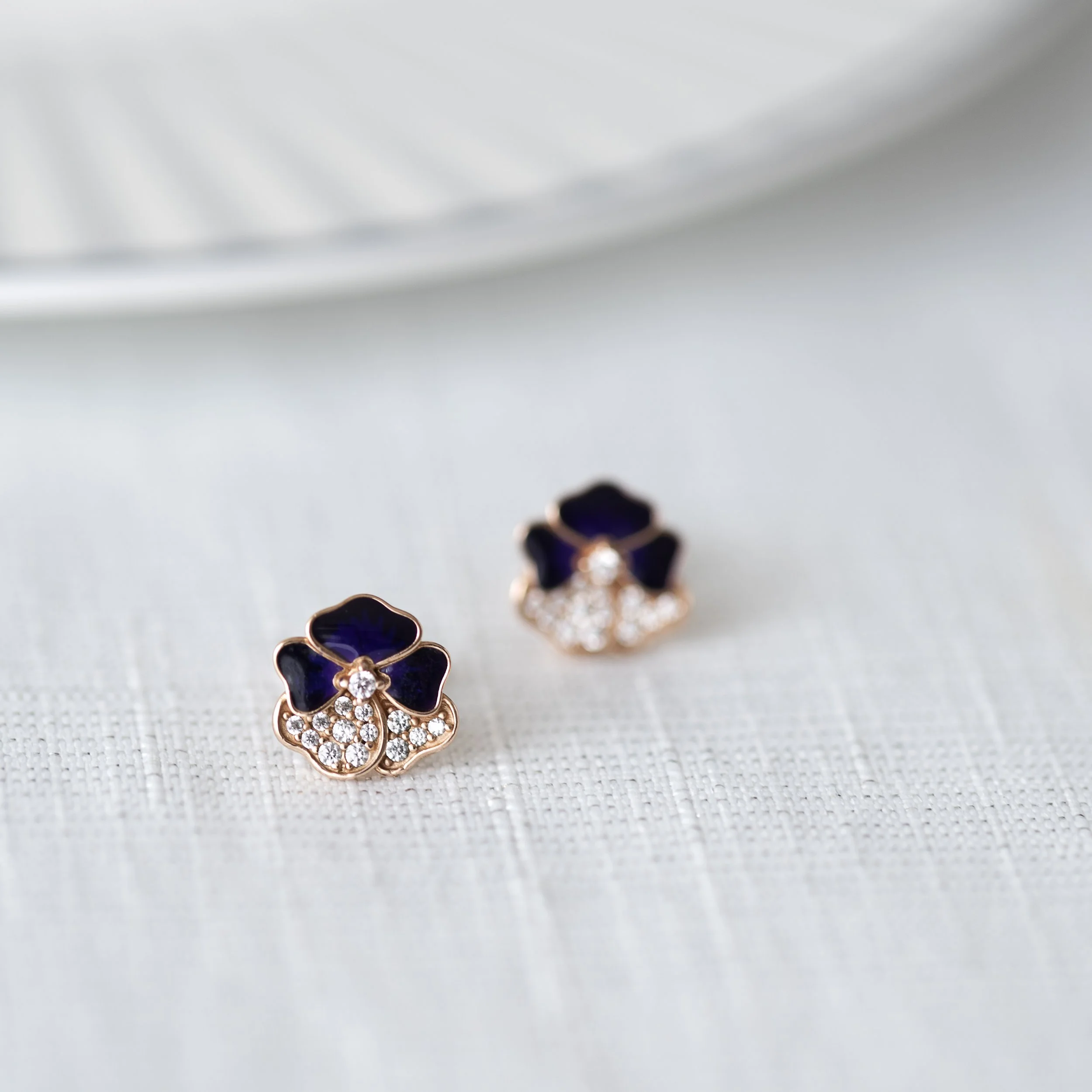 Pair of floral-shaped earrings with dark purple petals and sparkling diamonds on a white textured fabric surface.