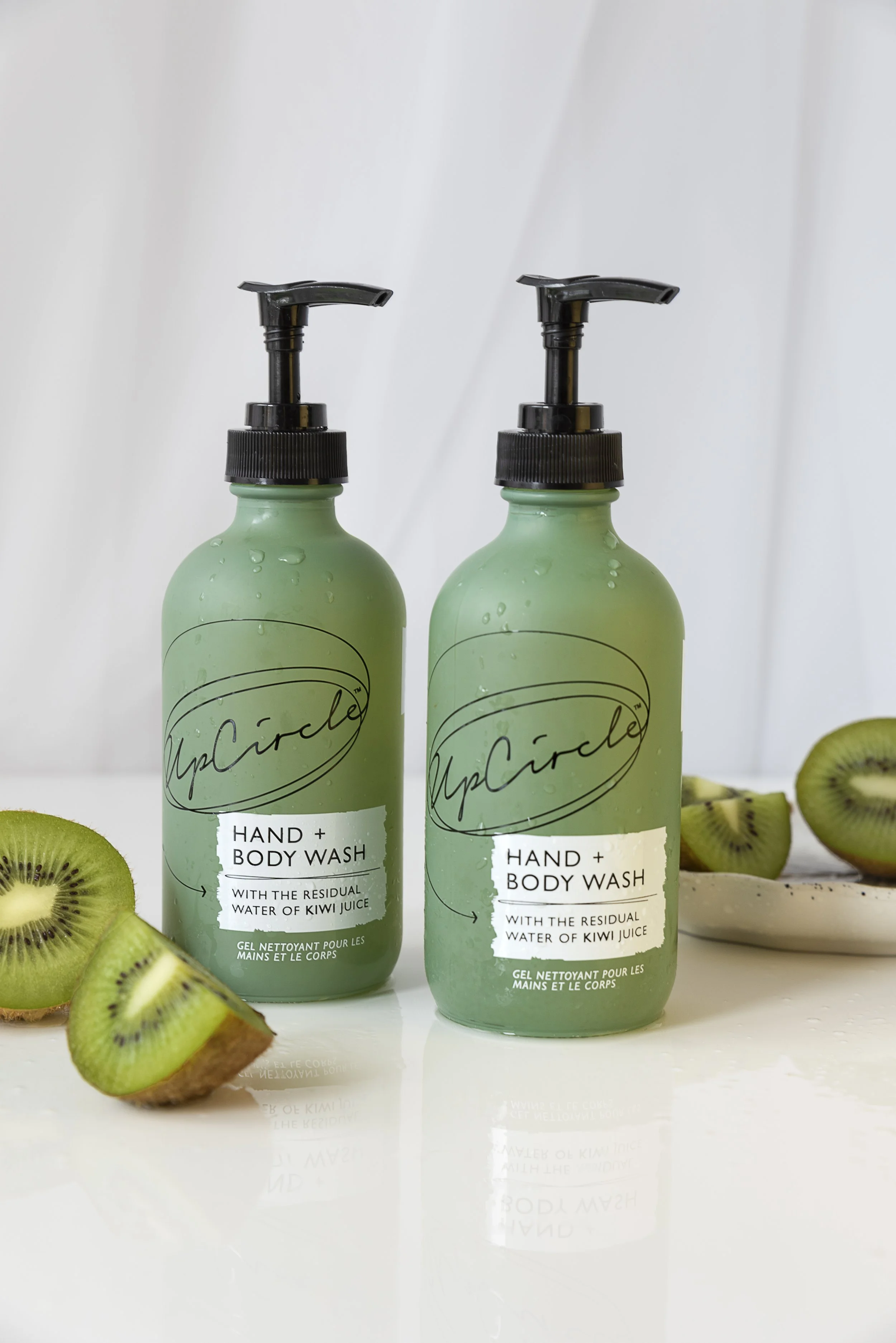 Two green bottles of hand and body wash with black pump dispensers, surrounded by sliced kiwifruit on a white surface.
