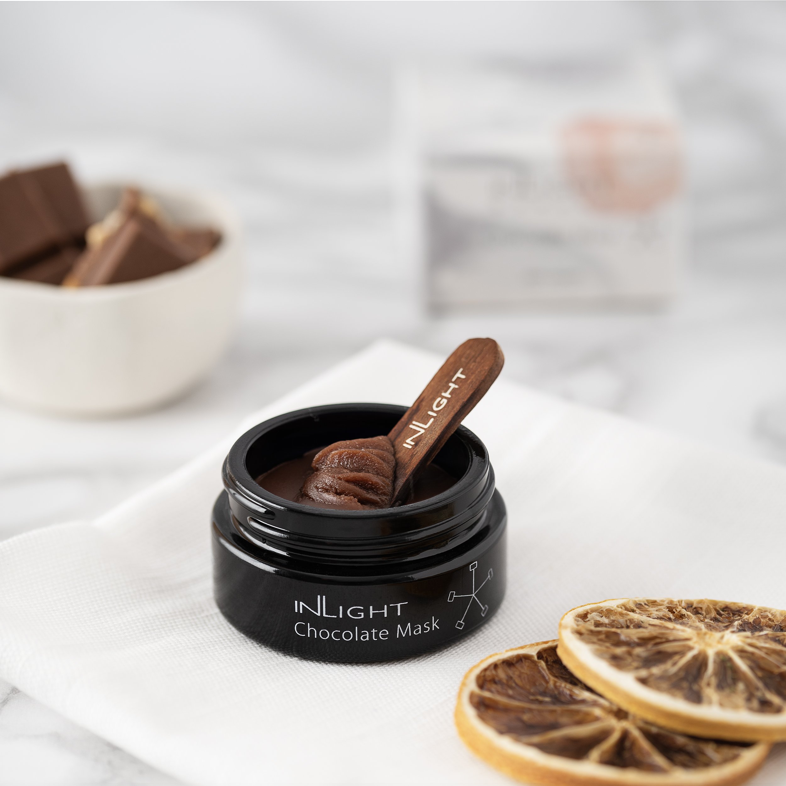 Open jar of INLight chocolate mask with a wooden spatula inside, placed on a white cloth. In the background, a bowl of chocolate pieces and slices of dried orange are visible.