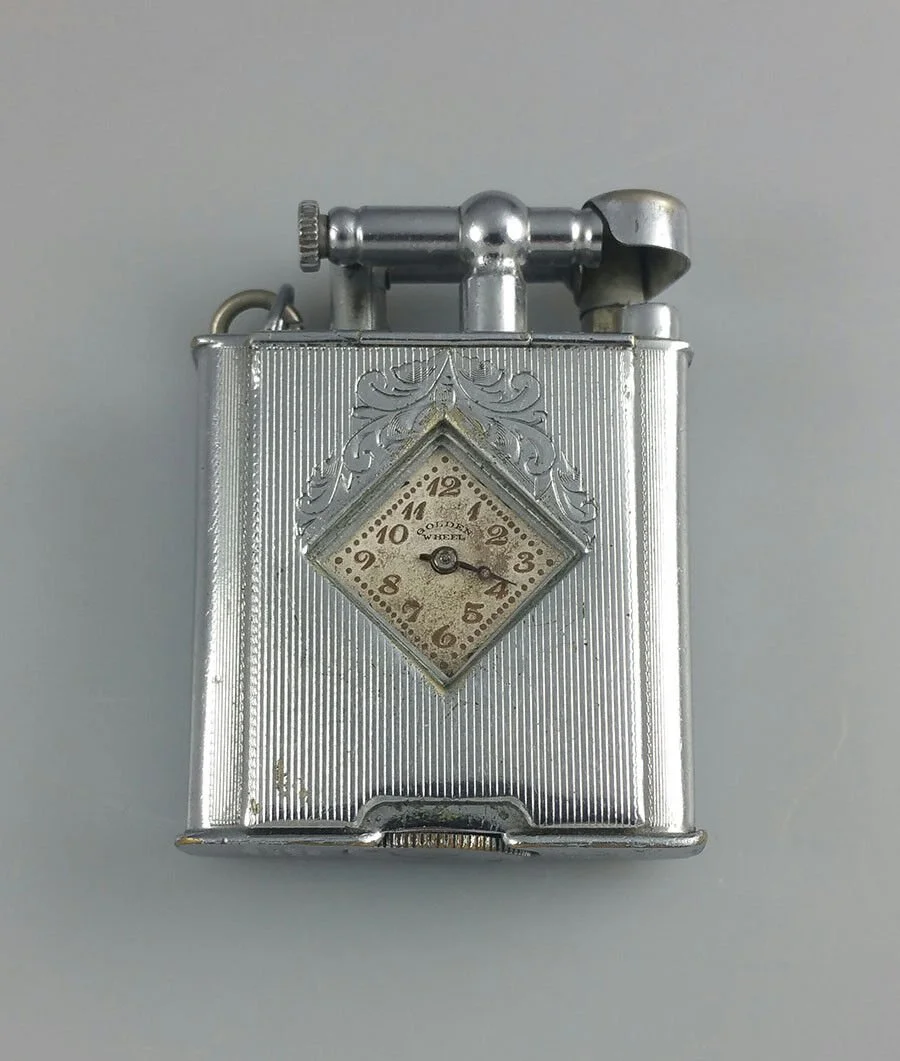A vintage silver cigarette lighter with a built-in clock on its front, featuring ornate design and roman numerals.