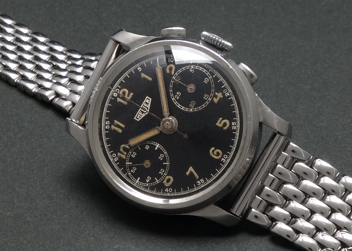 A vintage Heuer chronograph wristwatch with a black dial, gold Arabic numerals, and a silver metal bracelet.